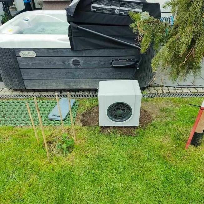 Zewnętrzny widok wanny z hydromasażem marki Wellis z częściowo otwartą pokrywą, obok zainstalowany biały panel sterujący, na trawniku z widocznymi elementami instalacji.