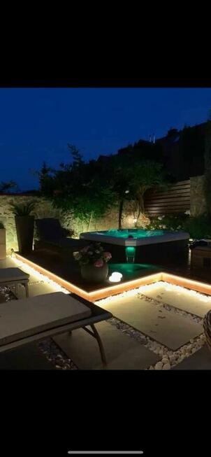 Oświetlona strefa relaksu z hydromasażem na zewnątrz, widok nocą, otoczona dekoracyjnym oświetleniem LED i kamienną ścieżką.