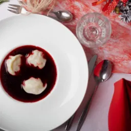 Wigilijny barszcz z uszkami w białym, głębokim talerzu, ustawiony na świątecznym stole z dekoracjami, widoczne sztućce, szklanka wody i logo firmy cateringowej w rogu zdjęcia.