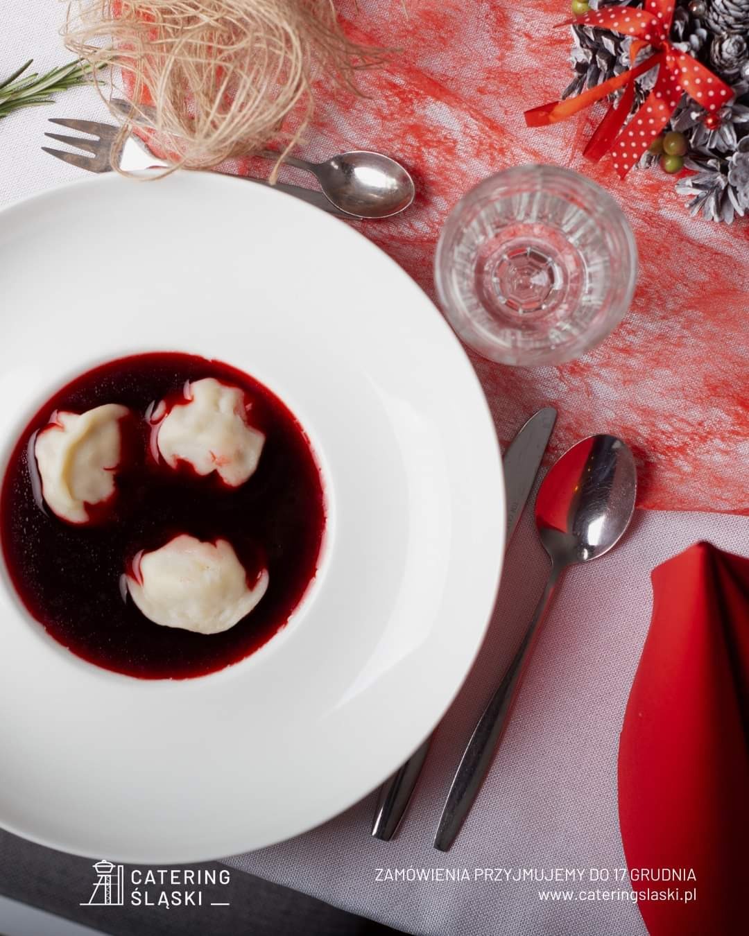 Wigilijny barszcz z uszkami w białym, głębokim talerzu, ustawiony na świątecznym stole z dekoracjami, widoczne sztućce, szklanka wody i logo firmy cateringowej w rogu zdjęcia.