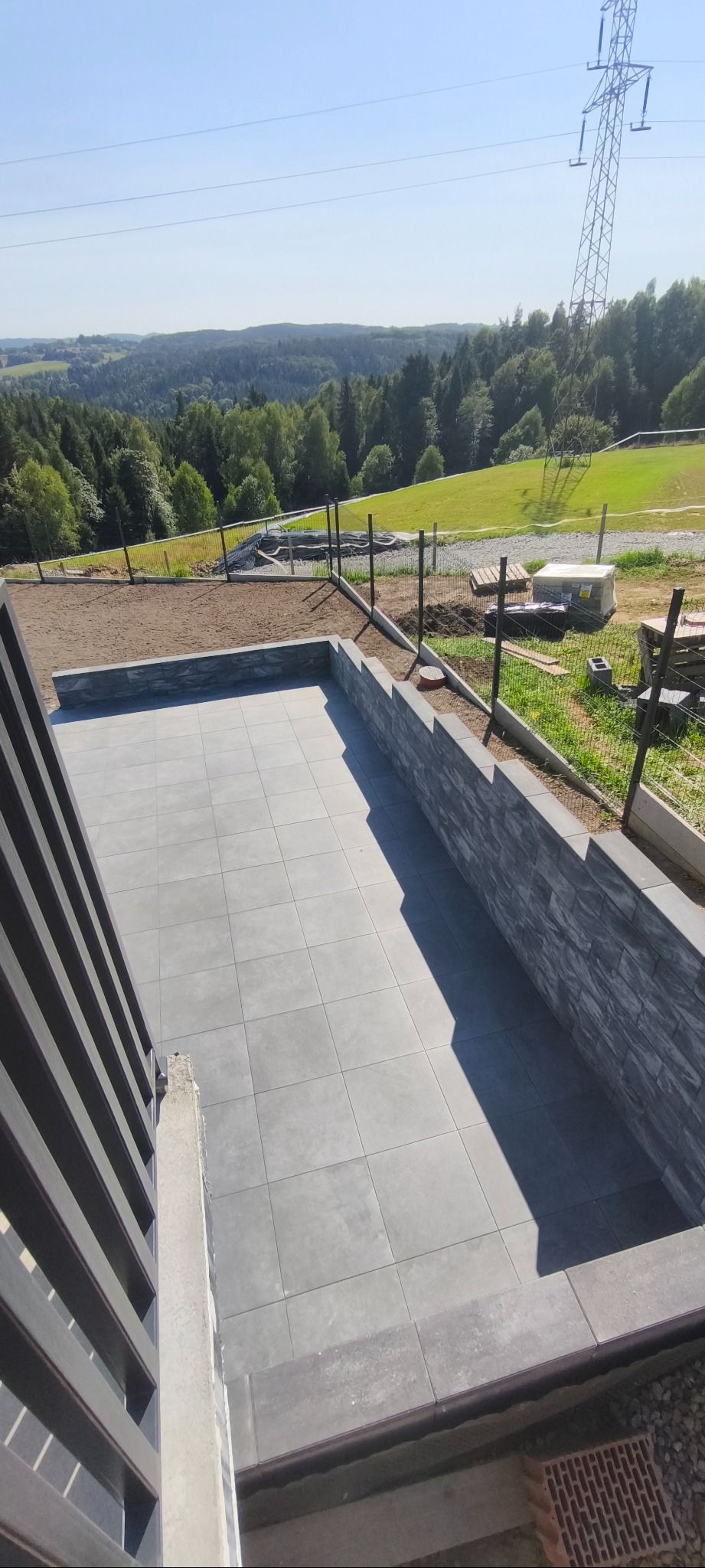 Szare płyty tarasowe ułożone na zewnątrz, otoczone murem z kamienia dekoracyjnego. W tle zielony krajobraz z lasem i słupem wysokiego napięcia.