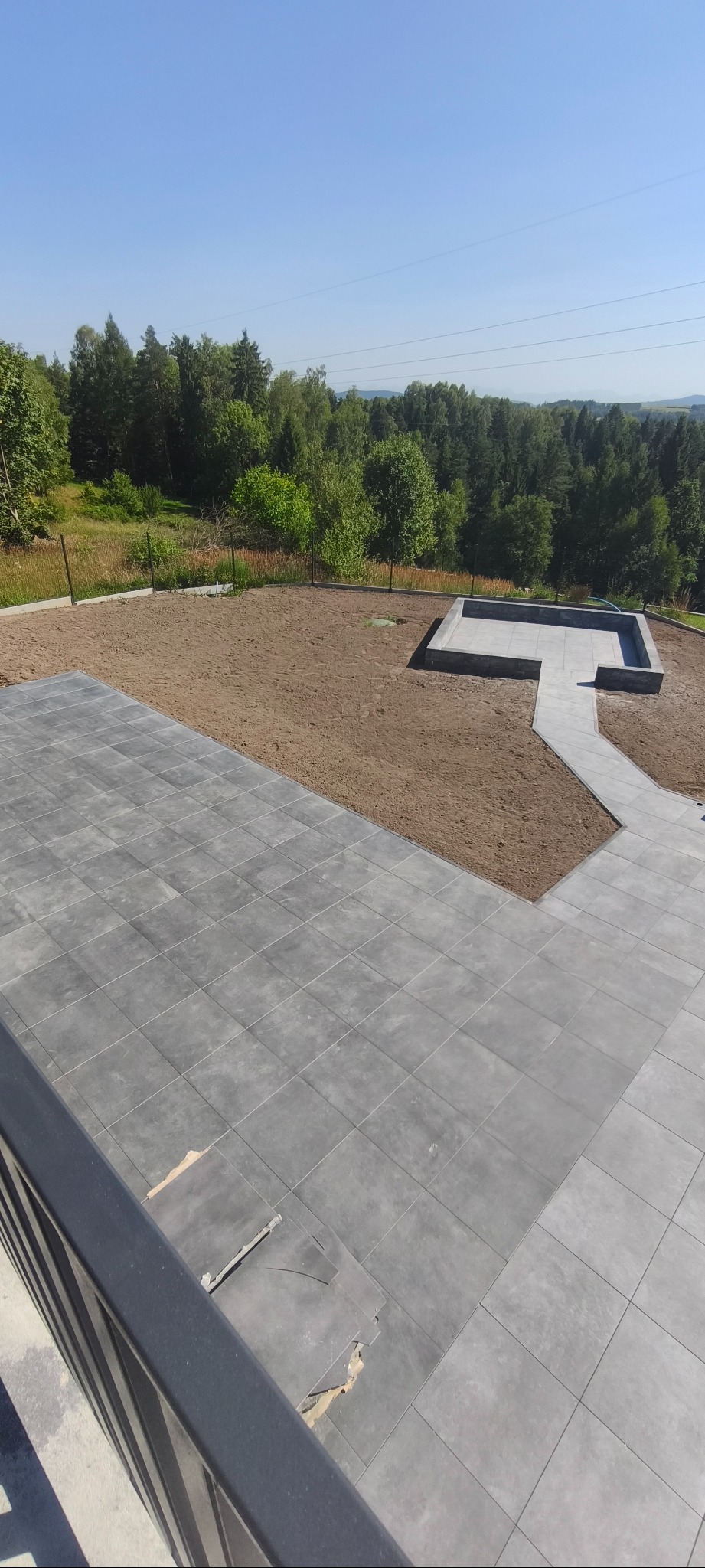 Widok z góry na szare płyty chodnikowe i przygotowany teren pod trawnik z obrzeżem, w tle zieleń drzew i błękitne niebo. Fragment uszkodzonej nawierzchni.