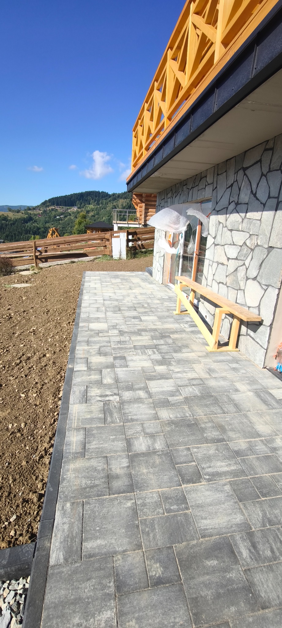 Szara kostka brukowa na tarasie domu z kamienną elewacją i drewnianą konstrukcją, z widokiem na wzgórza w tle. Częściowo przygotowany teren pod trawnik.