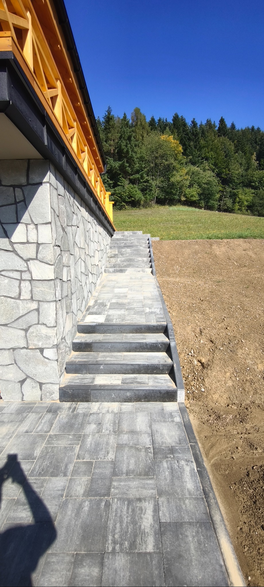 Ułożona kostka brukowa prowadząca do schodów przy budynku z kamienną elewacją. W tle drzewa i niebo. Ciemnoszare kostki tworzą wzór. Widoczny cień osoby robiącej zdjęcie.