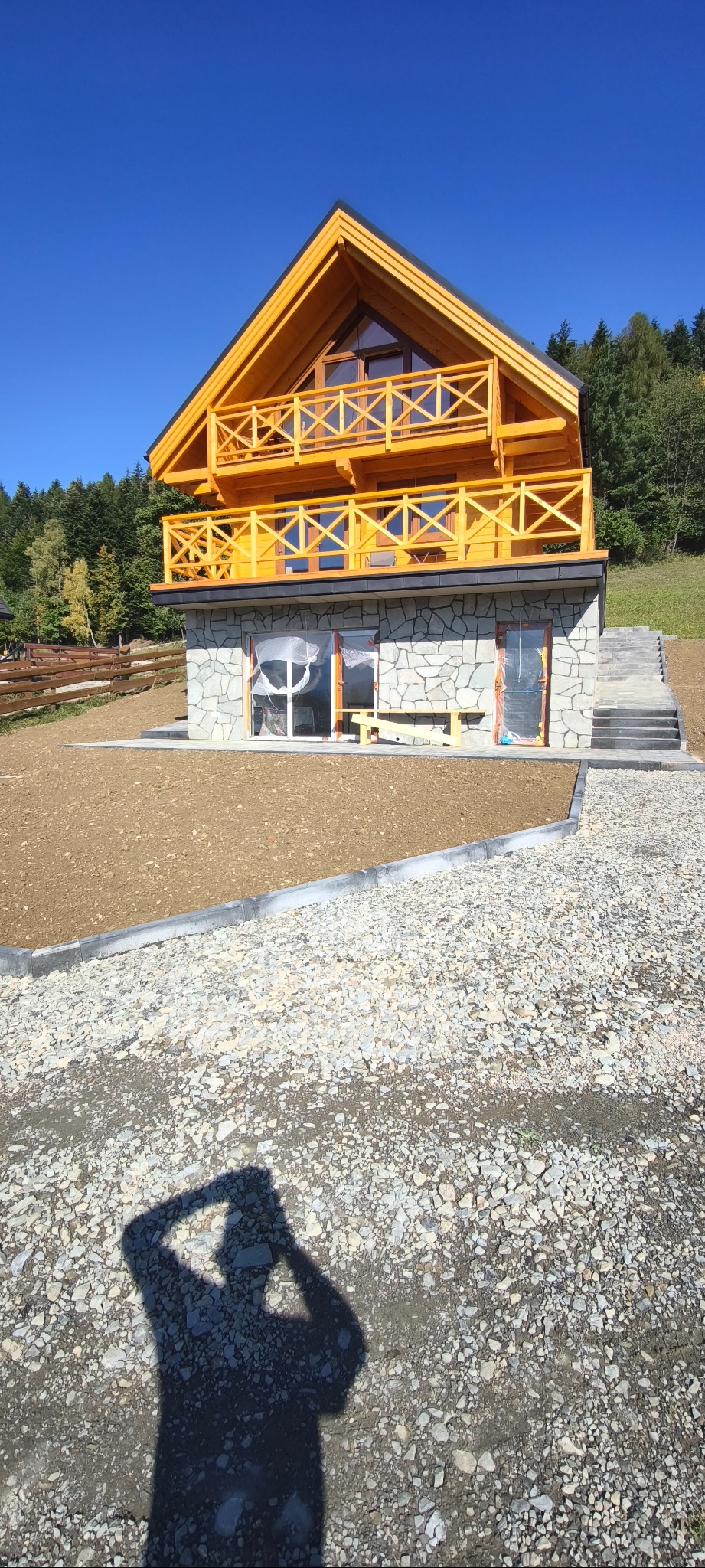 Nowy dom z drewnianą konstrukcją i kamienną elewacją, otoczony przygotowanym terenem i ścieżką z kamienia. Widoczny cień osoby robiącej zdjęcie.