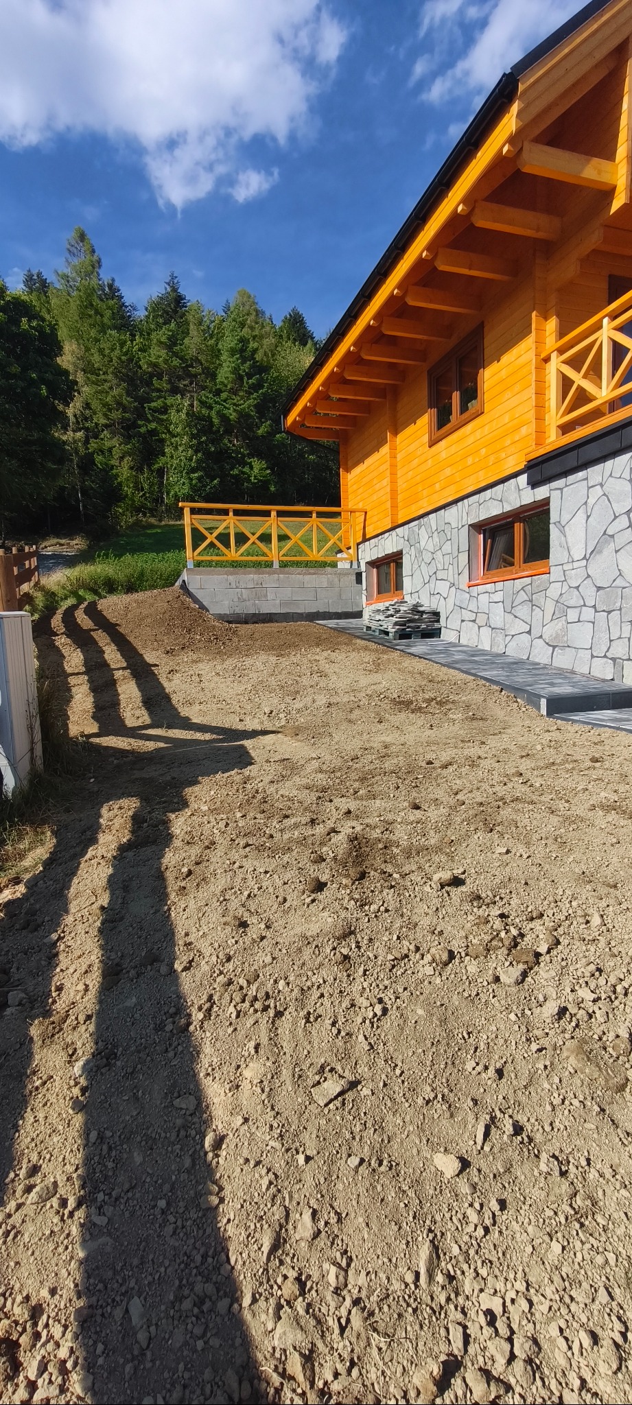 Przygotowany teren pod budowę lub zagospodarowanie działki, z widocznym fragmentem drewnianego domu i ułożonym chodnikiem z kostki. Widoczne ślady opon.