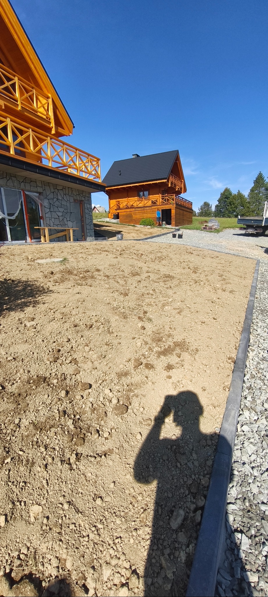 Przygotowany teren pod budowę, widoczne dwa drewniane domy w tle, jeden z kamienną elewacją. W tle niebieskie niebo. W rogu cień osoby robiącej zdjęcie.