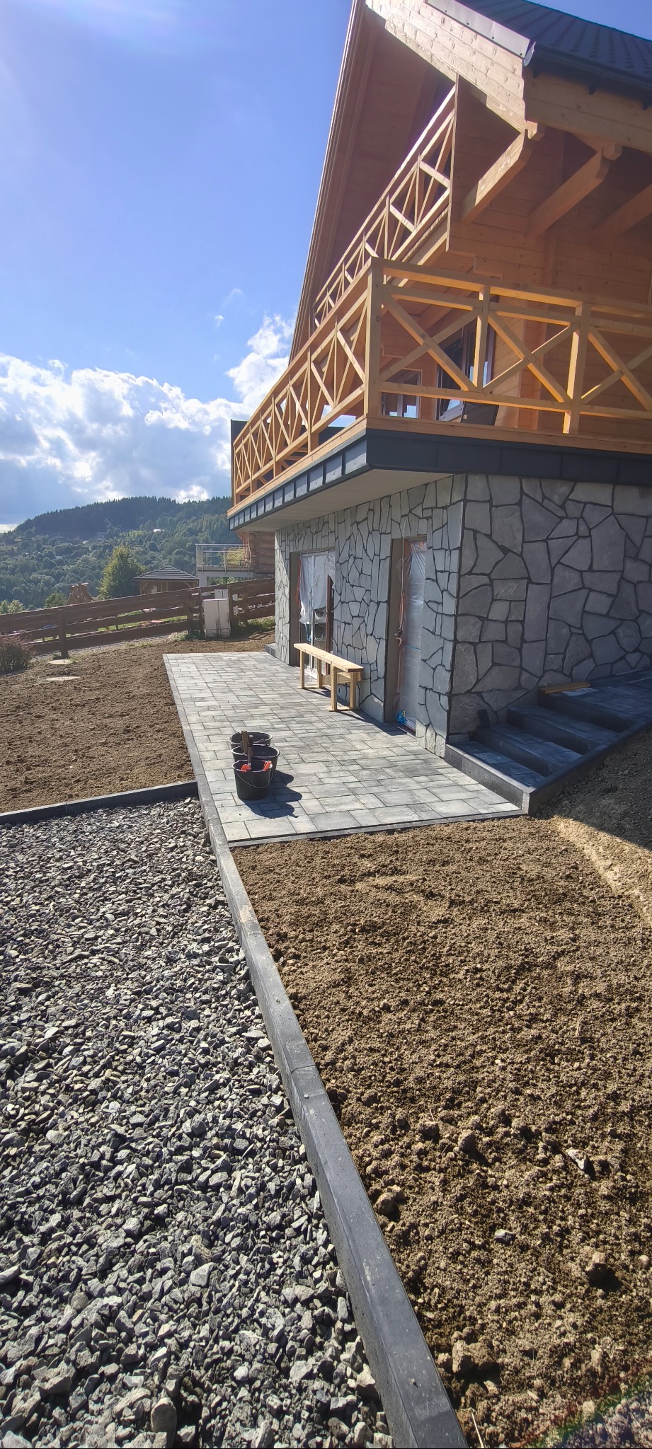 Ukończony taras z kostki brukowej przy domu z kamienną elewacją i drewnianym balkonem. Widoczne przygotowane rabaty, krajobraz górski w tle.