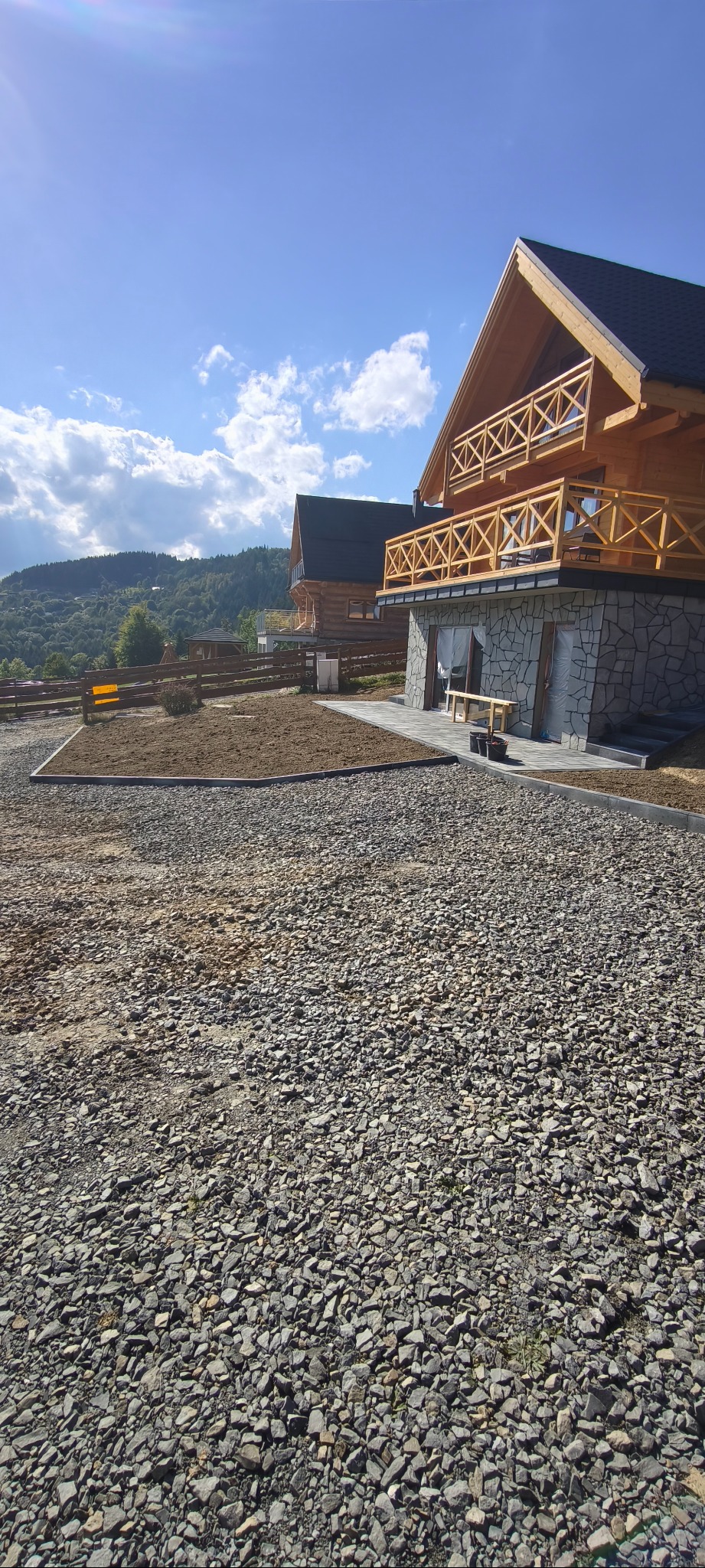 Dwa drewniane domy z kamiennymi elementami, teren przygotowany pod zagospodarowanie, wysypany kamieniem, obrzeża z kostki. W tle góry i błękitne niebo.