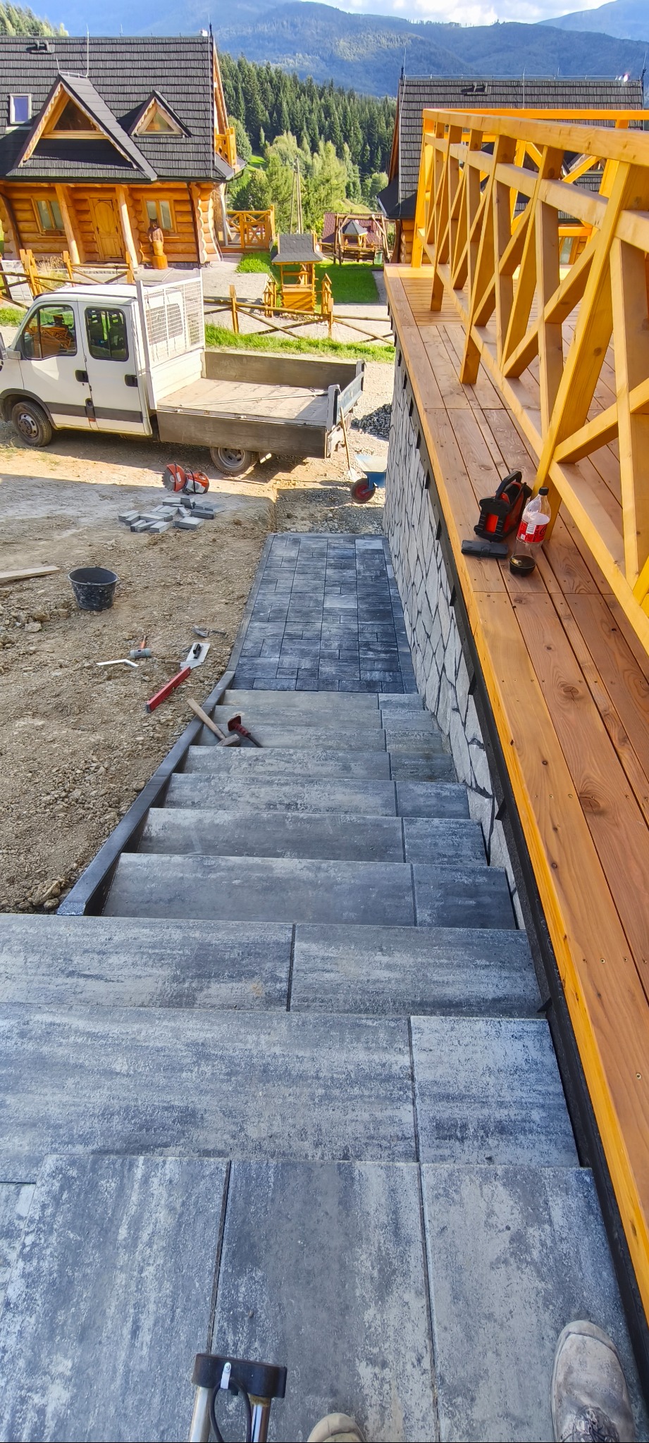 Schody z kostki brukowej w odcieniach szarości, prowadzące w dół obok drewnianego tarasu. W tle domki w stylu góralskim i góry. Widoczne narzędzia pracy.