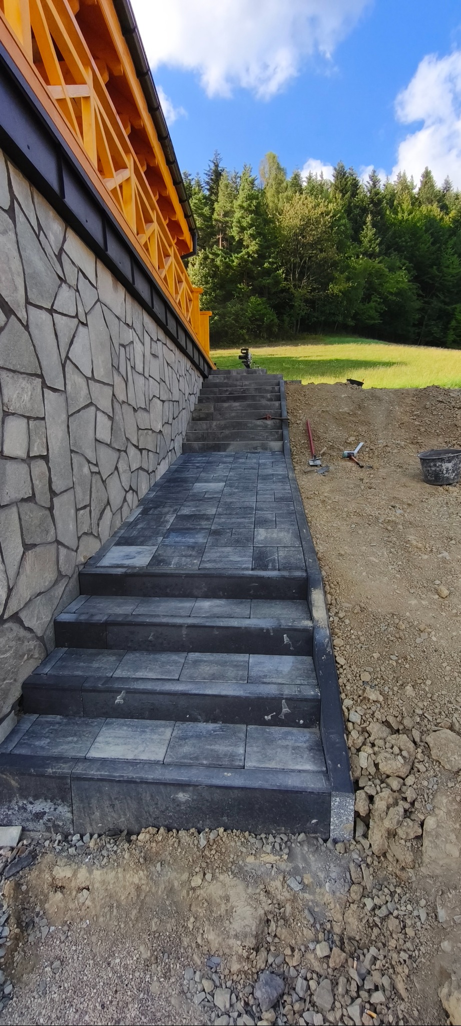 Schody z ciemnej kostki brukowej prowadzą do domu z kamienną elewacją i drewnianymi elementami. Widoczne narzędzia i teren w trakcie prac ziemnych.