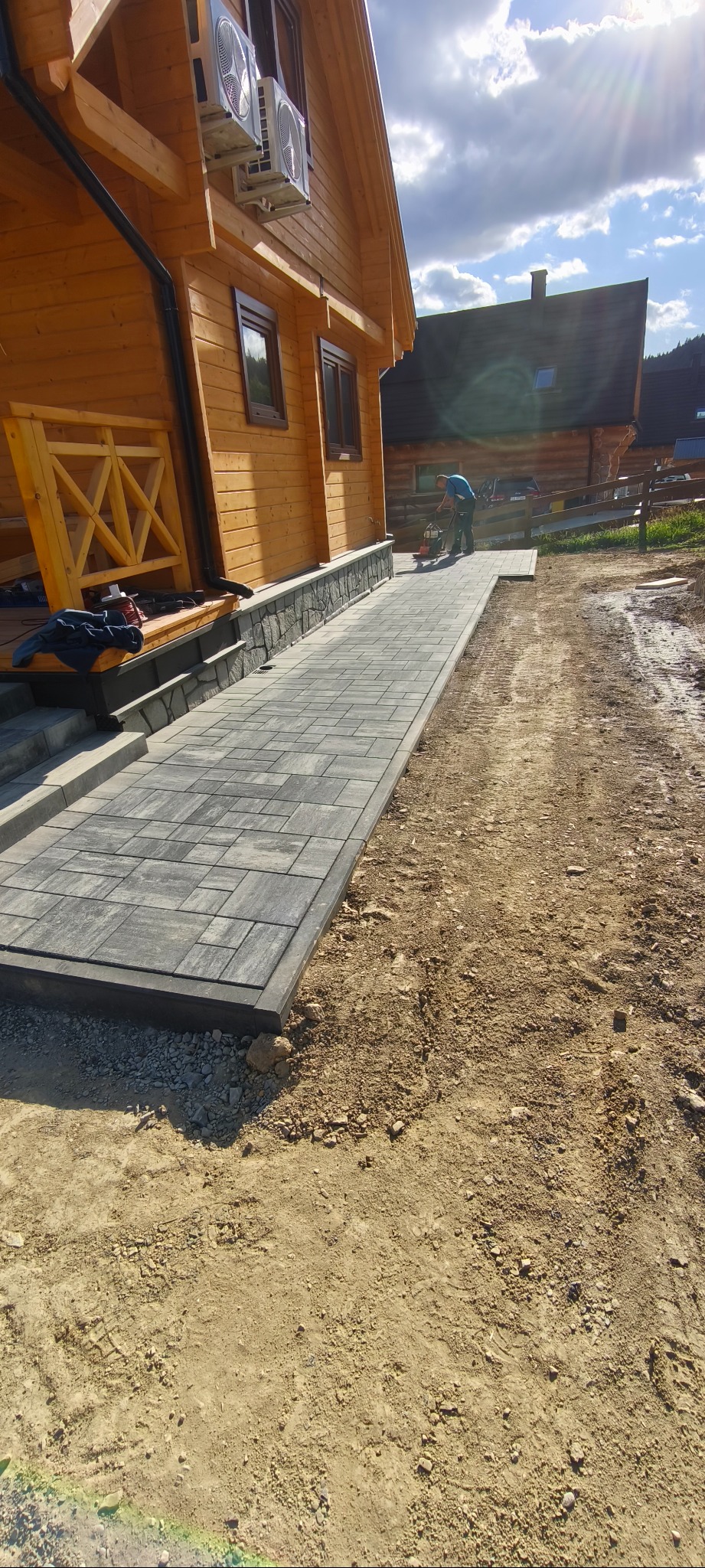 Ułożona, szara kostka brukowa prowadząca do drewnianego domu. W tle pracownik zagęszcza podłoże pod dalszą część chodnika. Widoczne nierówności terenu.