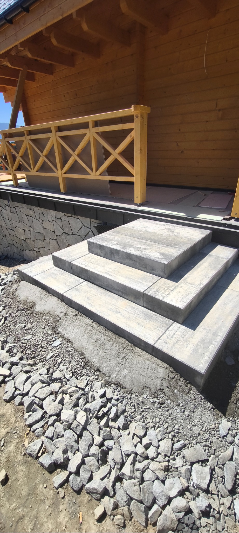 Schody z kostki brukowej prowadzące do drewnianego domu z balustradą, otoczone kamieniami. Widok z dołu, podkreślający solidność konstrukcji i detale wykończenia.