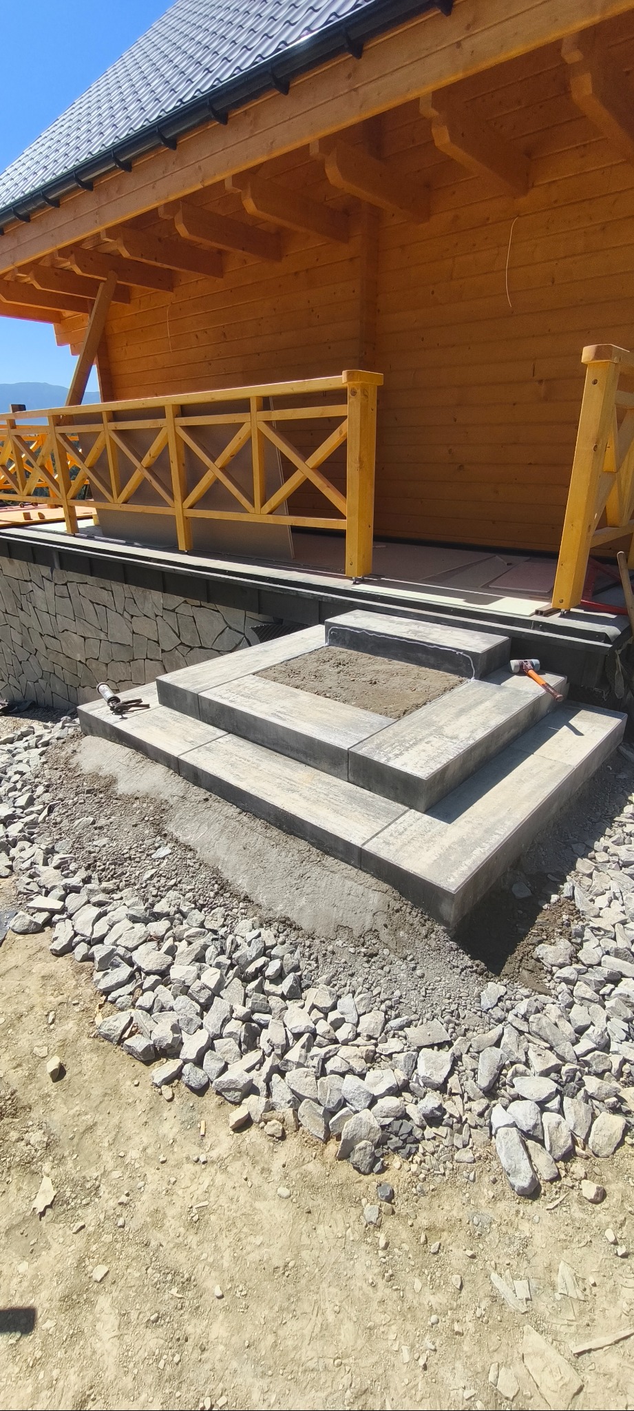 Schody z kostki brukowej prowadzące do drewnianego domu, częściowo wykończone, z widocznymi narzędziami pracy i kamieniami w tle.