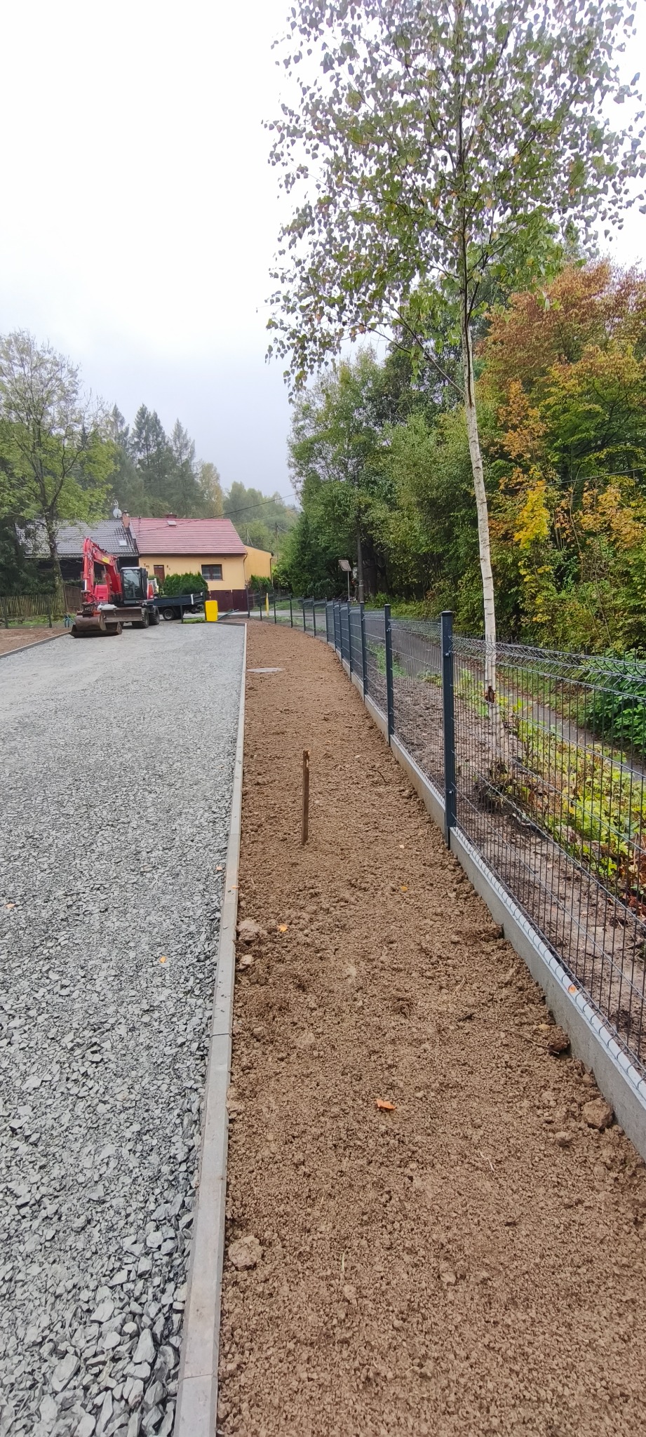 Teren po niwelacji z przygotowanym podłożem pod kostkę brukową i ogrodzeniem panelowym. W tle widoczny sprzęt budowlany i zabudowania.