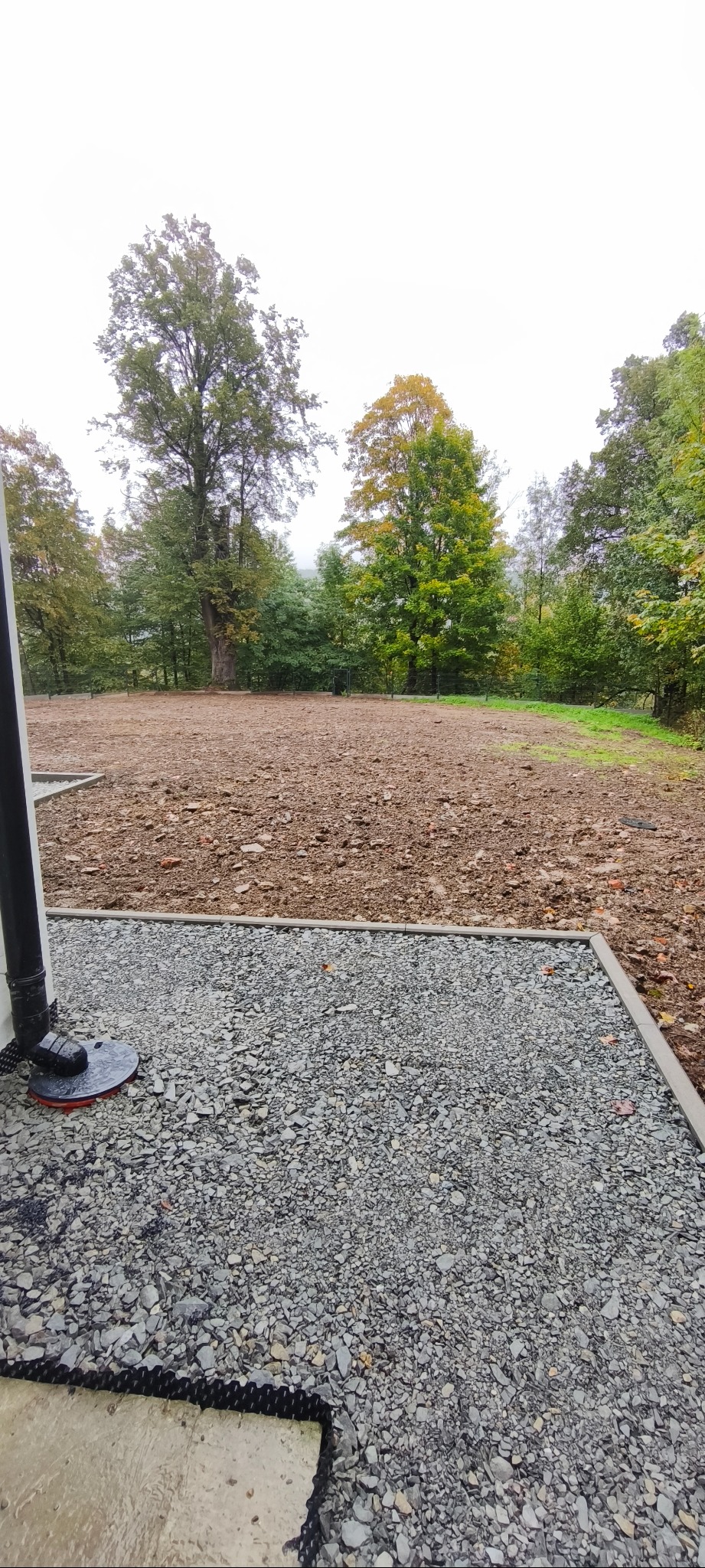 Przygotowany teren pod budowę z częściowo wyłożonym szarym żwirem i widoczną rurą spustową, otoczony drzewami w jesiennej scenerii.