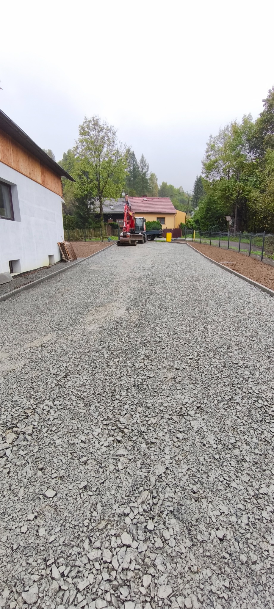 Szeroki plan utwardzonej drogi z szarego kruszywa, obrzeża betonowe, w tle minikoparka i budynki. Widok na posesję po niwelacji terenu i przygotowaniu podłoża.