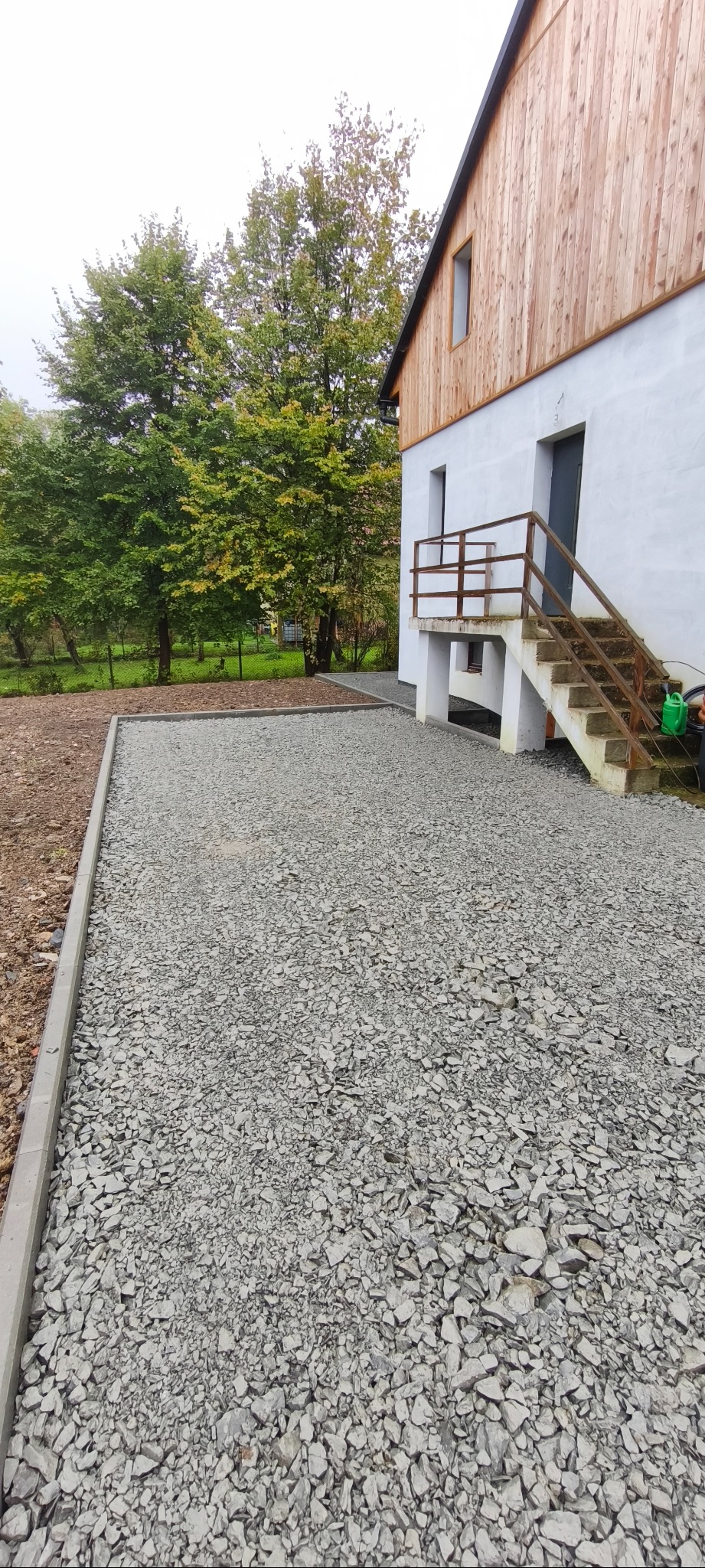 Utwardzony teren szarą, drobną frakcją kamienia, obrzeża wykończone betonową kostką. W tle budynek z drewnianą elewacją i zielone drzewa.