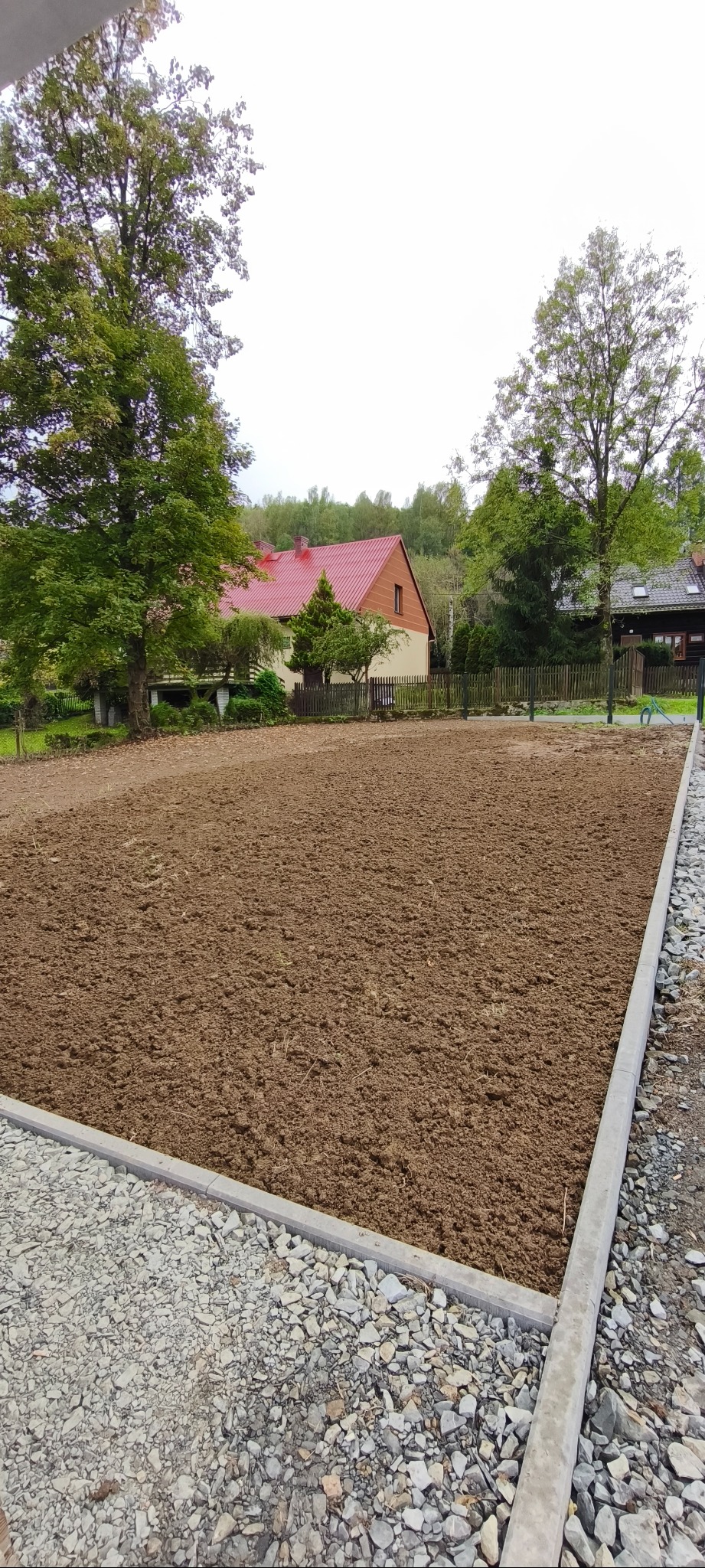 Przygotowany teren pod budowę, wyrównana ziemia otoczona betonową obwódką i kamieniami, w tle budynki mieszkalne i drzewa. Widok z poziomu gruntu.