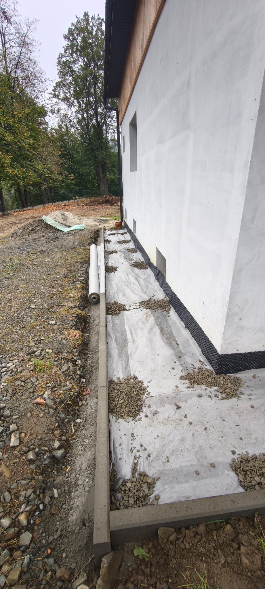 Przygotowanie terenu przy ścianie budynku: ułożona geowłóknina, obrzeża betonowe i rozsypany żwir. Widoczne fundamenty i fragment zieleni w tle.