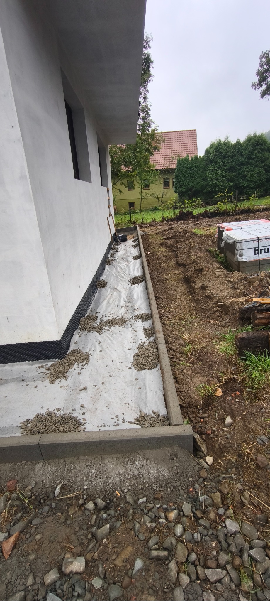 Przygotowanie podłoża pod kostkę brukową przy budynku: geowłóknina, drenaż z kamyków, obrzeża betonowe. Kostka brukowa w tle, zielone otoczenie.