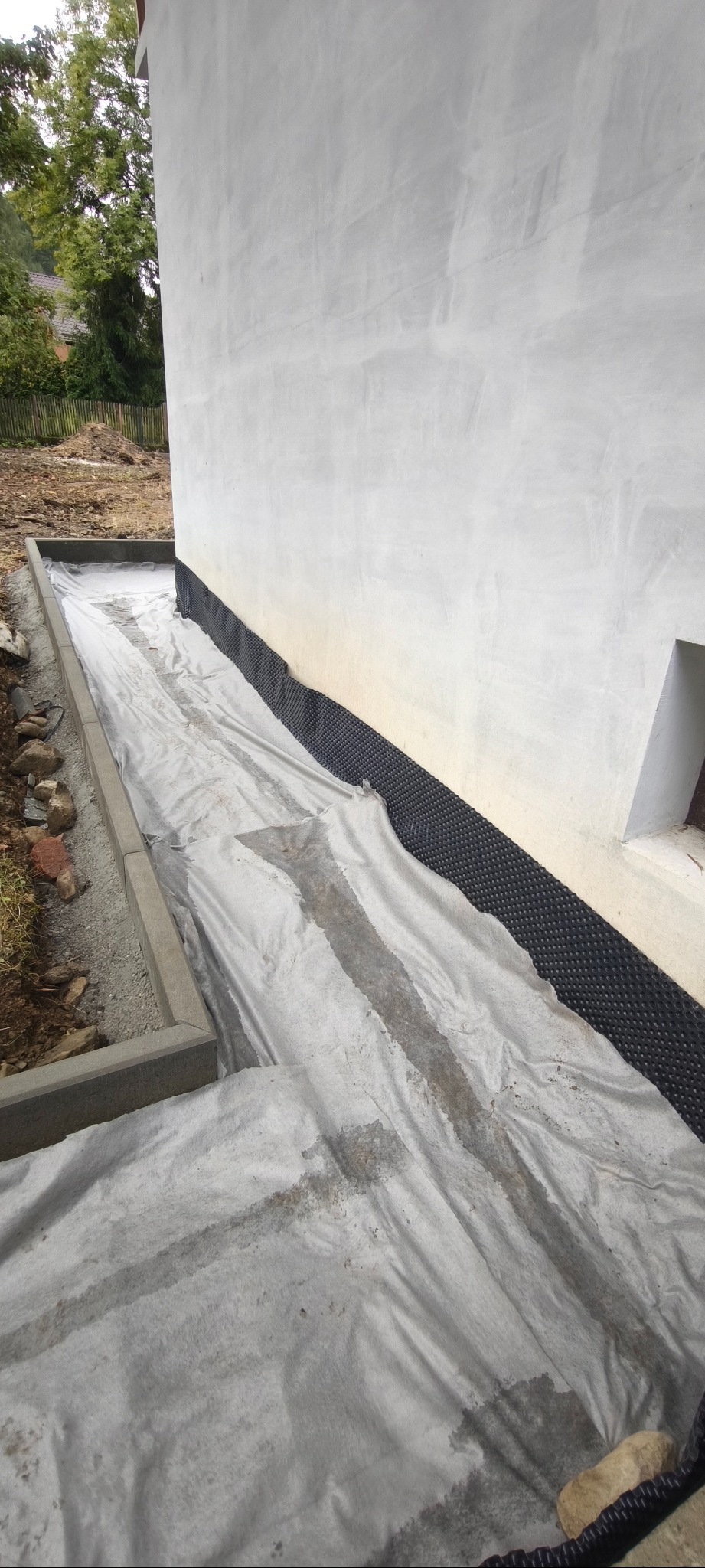 Przygotowanie terenu pod kostkę: obrzeża betonowe, geowłóknina i folia kubełkowa przy ścianie budynku. Widok na fragment ogrodu.