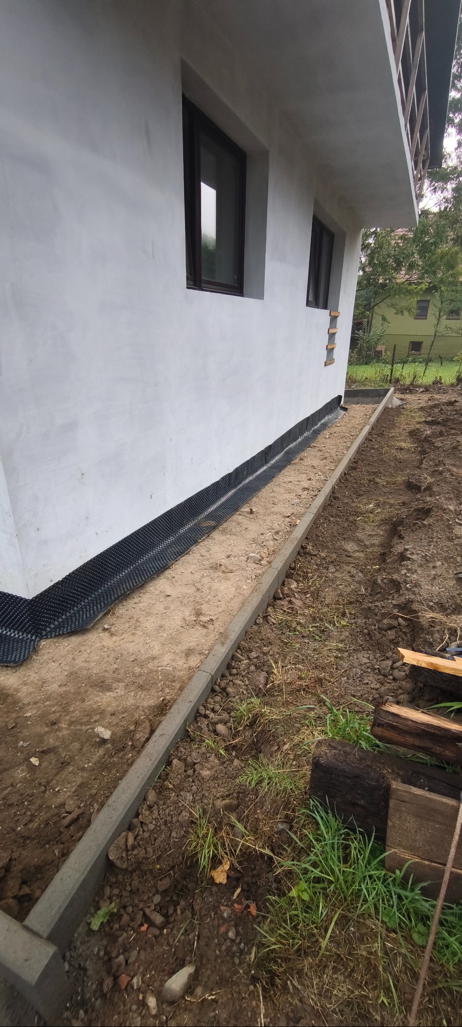 Fundament budynku z izolacją pionową i obrzeżami chodnikowymi. Widoczne okna i fragment ogrodu w tle. Ujęcie z poziomu gruntu.