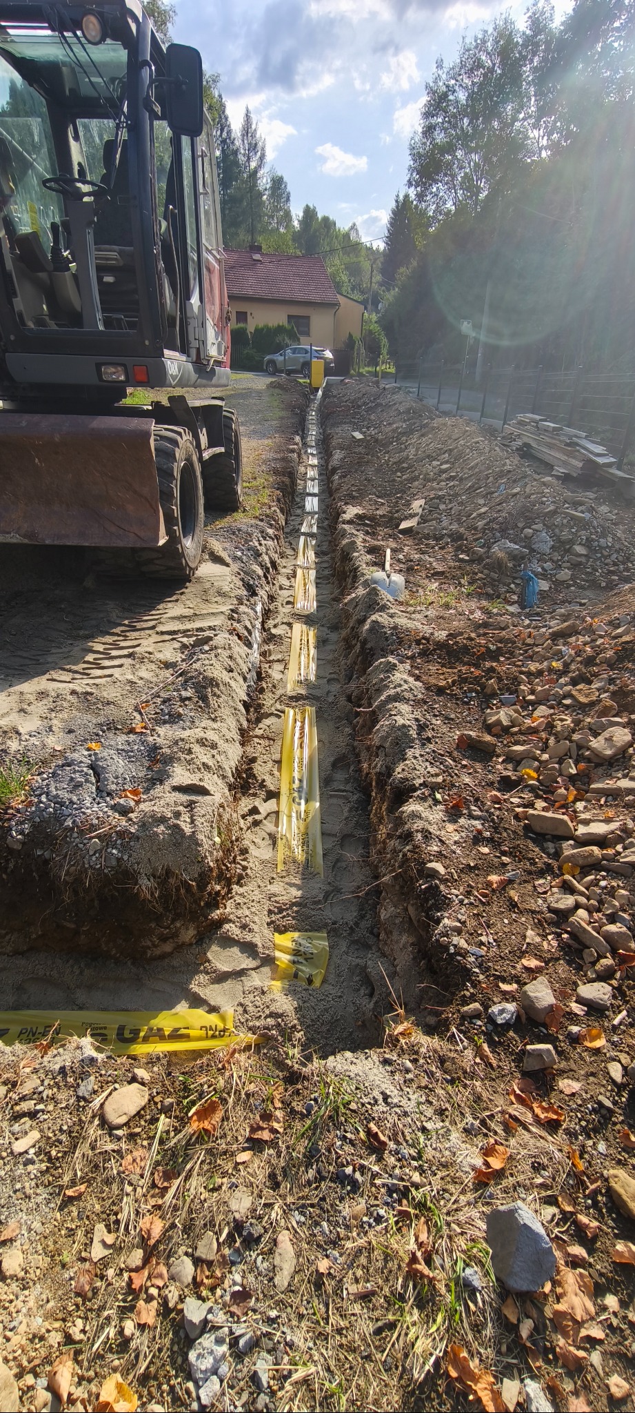 Wykop rowu pod instalacje gazowe, zabezpieczony żółtą folią ostrzegawczą. Na pierwszym planie gleba z liśćmi, w tle budynek i częściowo widoczna koparka.