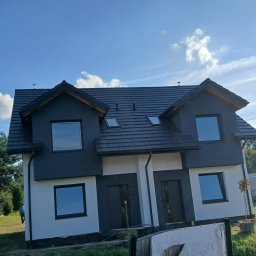 Daniel Leksander - Dwukondygnacyjny dom bliźniak z ciemną elewacją i dachem, jasnymi ścianami, okna z ciemnymi ramami, widok z zewnątrz w słoneczny dzień, drzewa w tle.