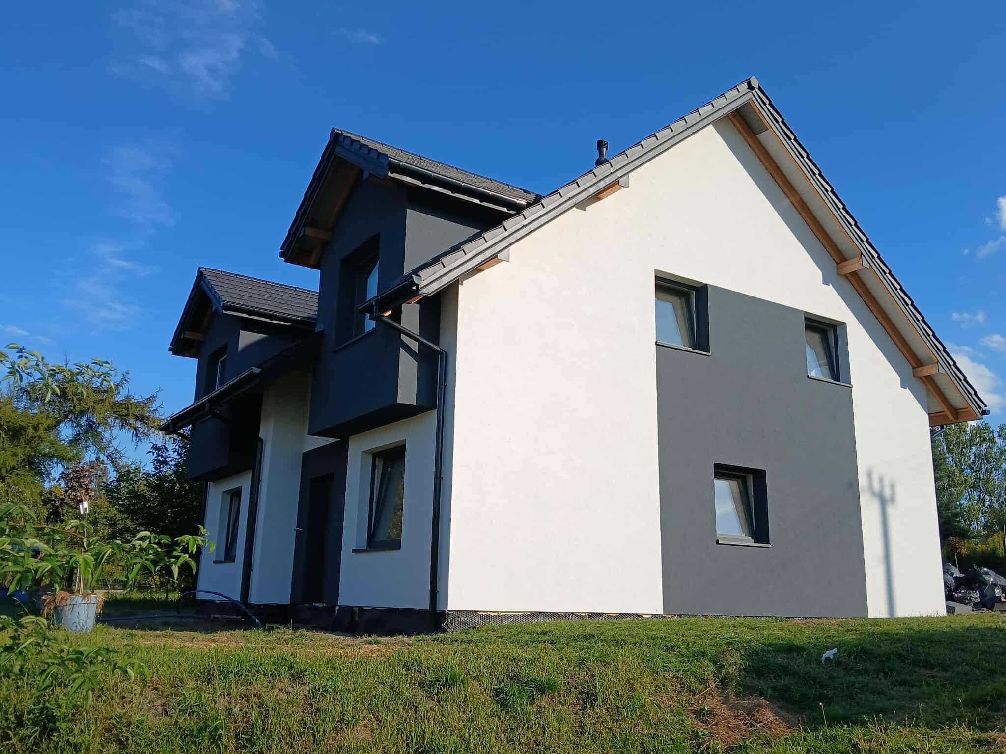 Nowoczesny dom z dwukolorową elewacją: białe i grafitowe ściany, ciemna dachówka, okna z grafitowymi ramami, na tle błękitnego nieba.