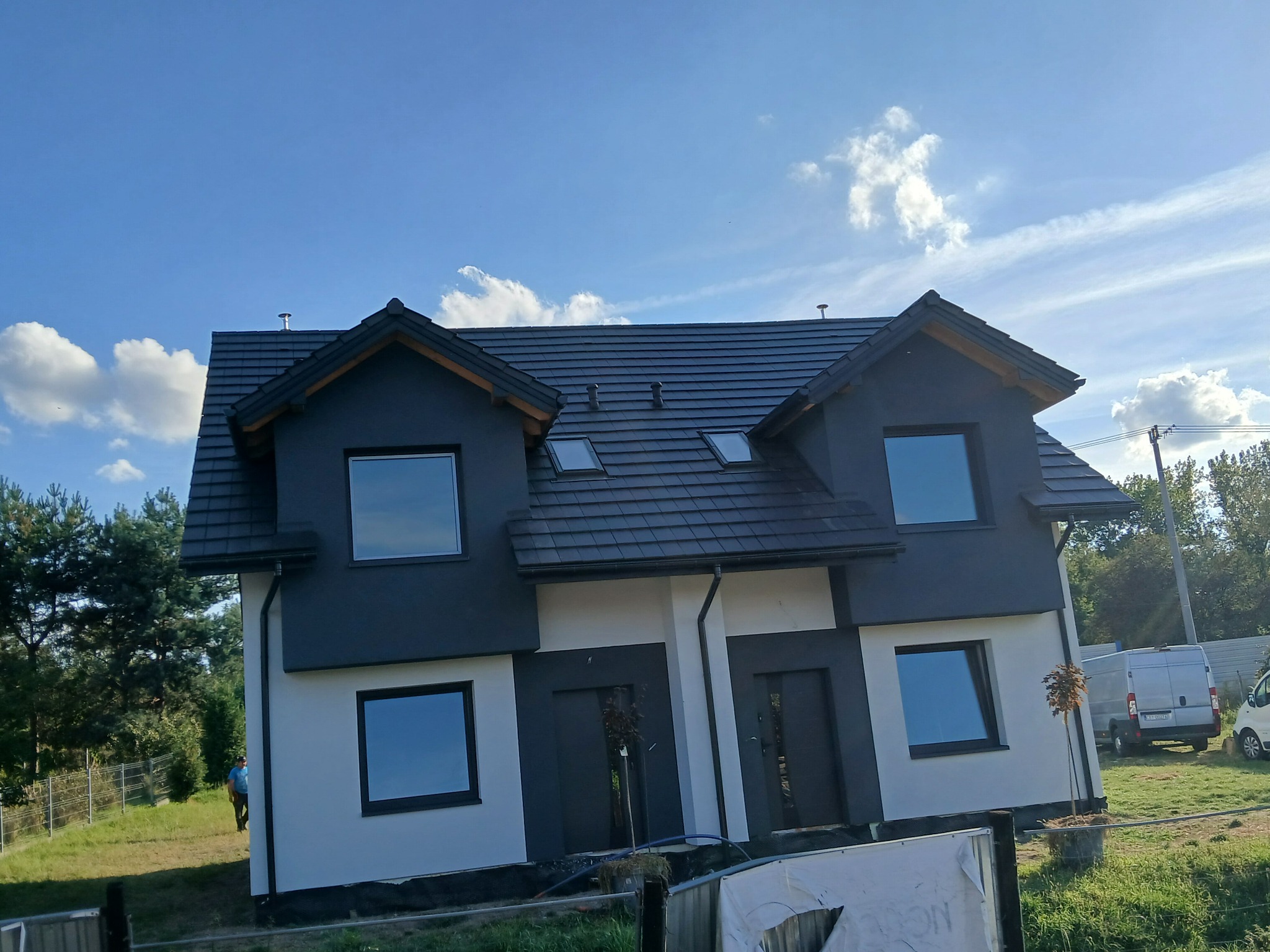 Dwukondygnacyjny dom bliźniak z ciemną elewacją i dachem, jasnymi ścianami, okna z ciemnymi ramami, widok z zewnątrz w słoneczny dzień, drzewa w tle.