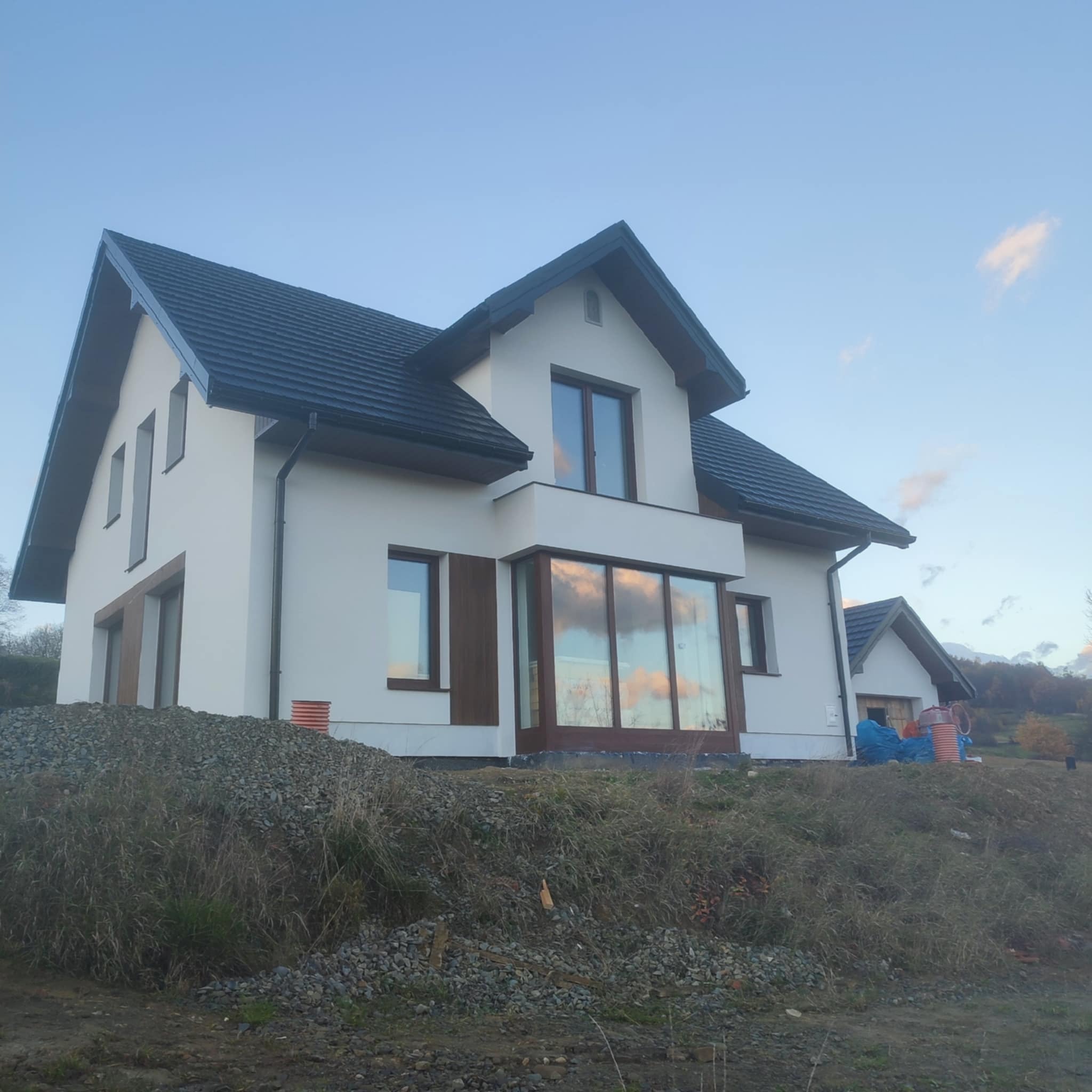 Nowoczesny dom jednorodzinny z białymi ścianami i ciemną dachówką, widziany z dołu, z hałdą gruzu na pierwszym planie. Okna z brązowymi ramami odbijają niebo.