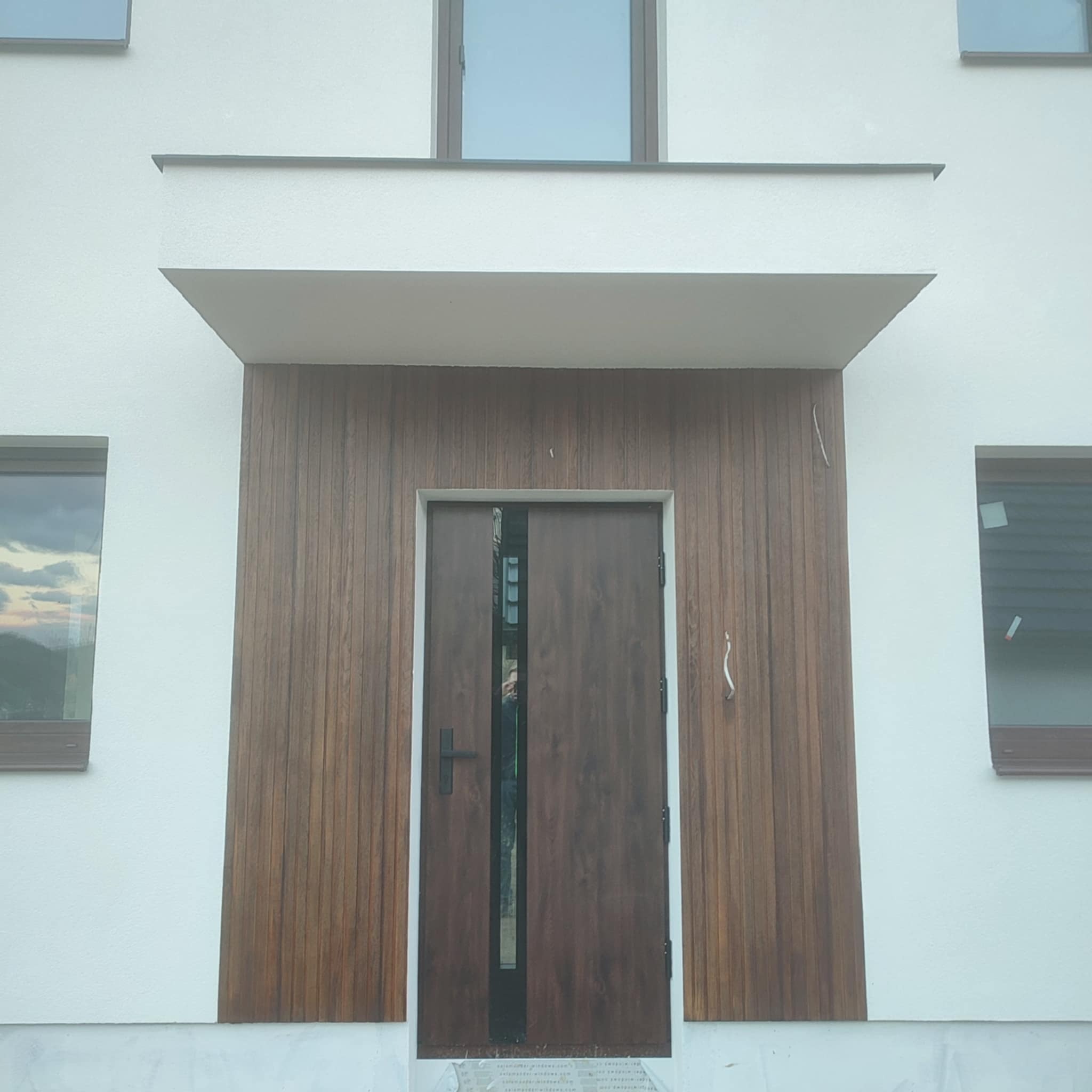 Nowoczesna elewacja domu z drewnianą okładziną wokół brązowych drzwi wejściowych i białym tynkiem. Widoczne okna z brązowymi ramami.