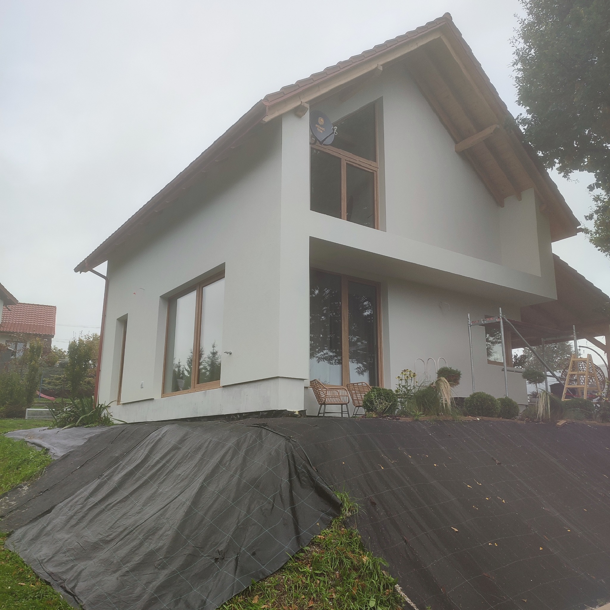 Nowoczesny dom jednorodzinny z jasną elewacją i drewnianymi oknami, widok z dołu na tle pochmurnego nieba. Na pierwszym planie zabezpieczona skarpa.