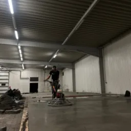 Pracownik w masce i słuchawkach wyrównuje mokrą, betonową posadzkę przemysłową za pomocą zacieraczki mechanicznej w hali z blachy falistej.