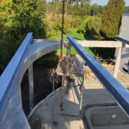 Budowa domu w stanie surowym otwartym, widoczne betonowe elementy konstrukcyjne i stalowe belki, rusztowanie robocze oraz otoczenie z zielenią.