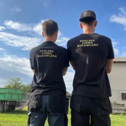 Dwóch mężczyzn w czarnych koszulkach z napisem 'Opolska Firma Budowlana' stoi tyłem, jeden w czapce z daszkiem, drugi w spodniach roboczych z narzędziami, na tle zielonego trawnika, przyczepy...