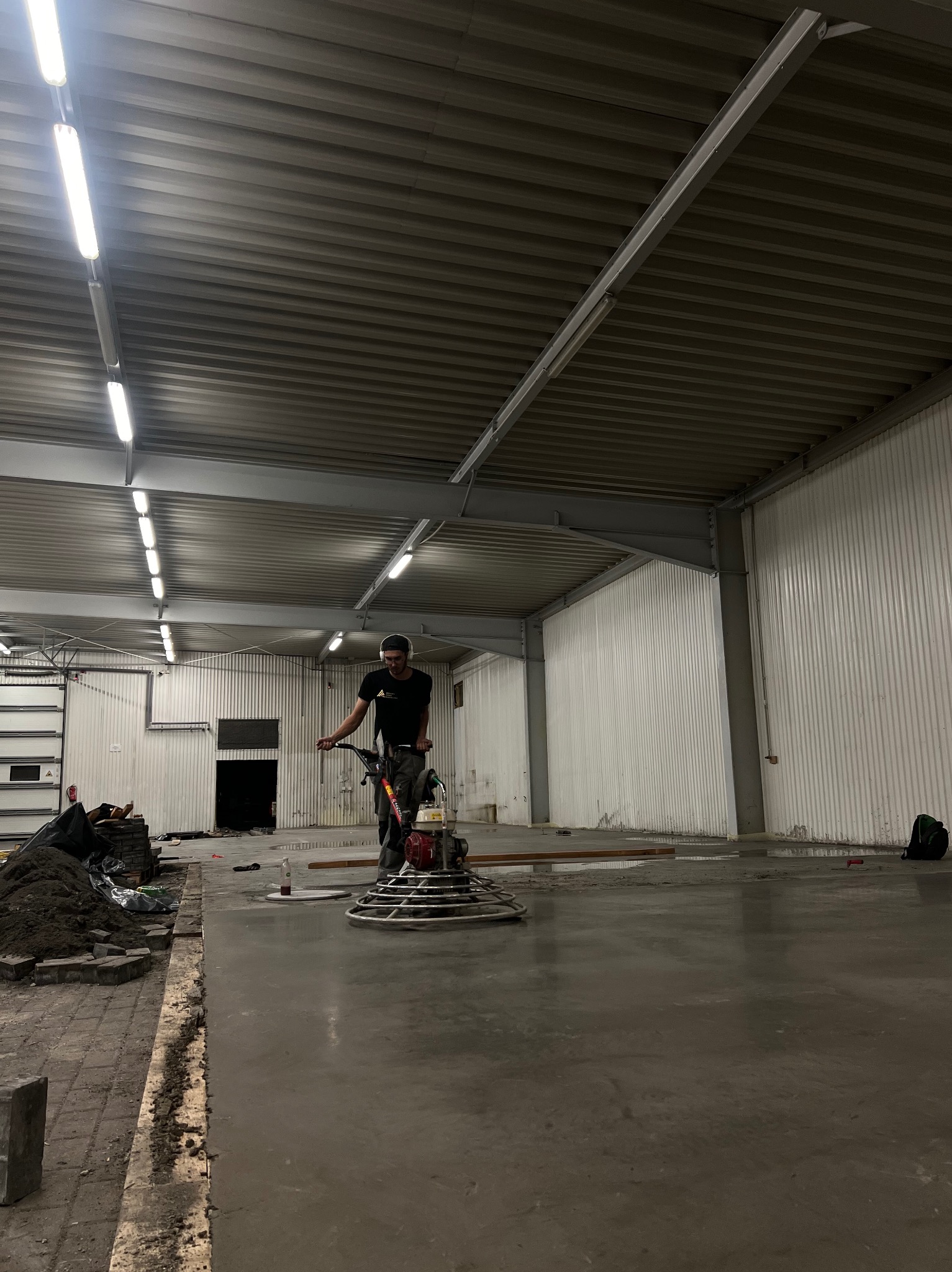 Pracownik w masce i słuchawkach wyrównuje mokrą, betonową posadzkę przemysłową za pomocą zacieraczki mechanicznej w hali z blachy falistej.