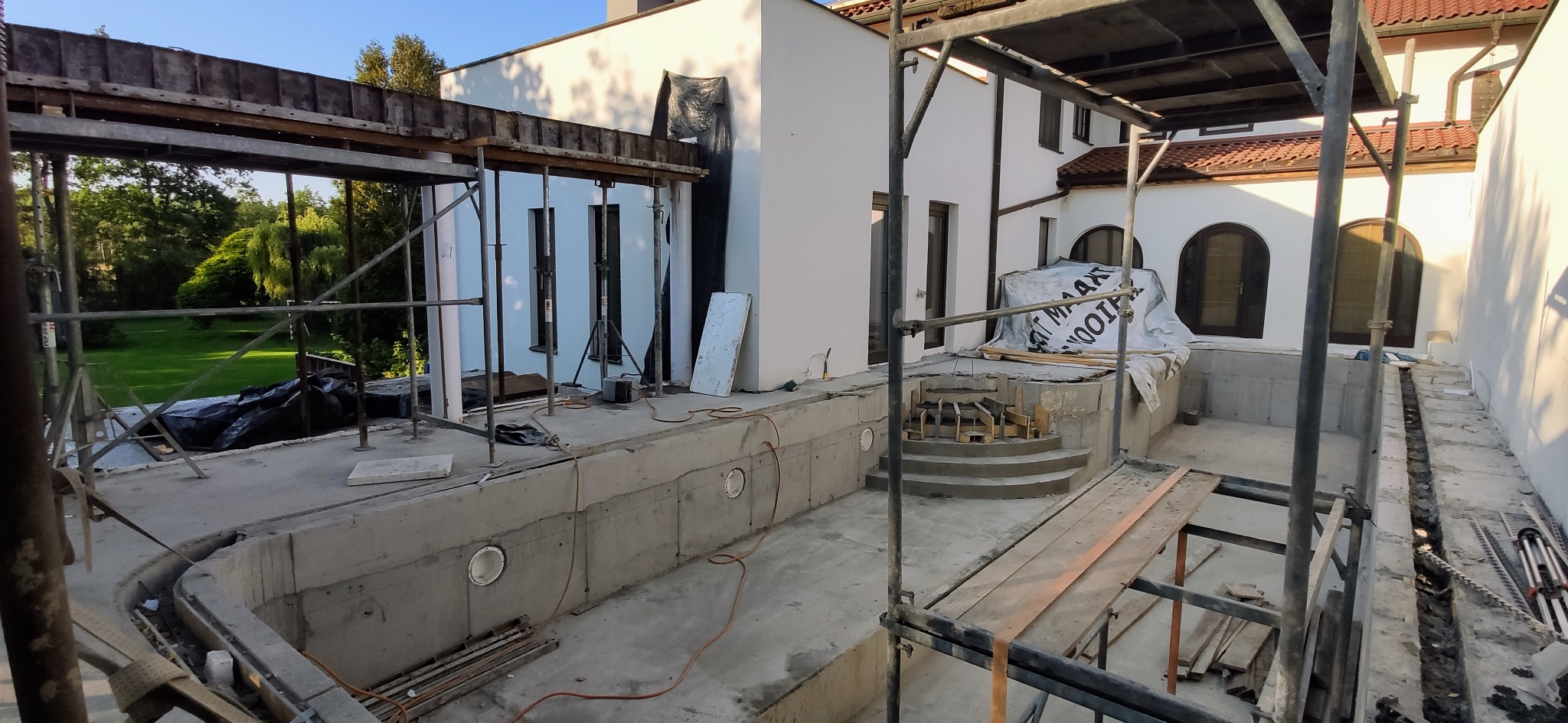 Betonowa konstrukcja niecki basenowej w trakcie budowy, widoczne szalunki, zbrojenia i stopnie wejściowe, na tle białego budynku z brązową dachówką i zielenią ogrodu.
