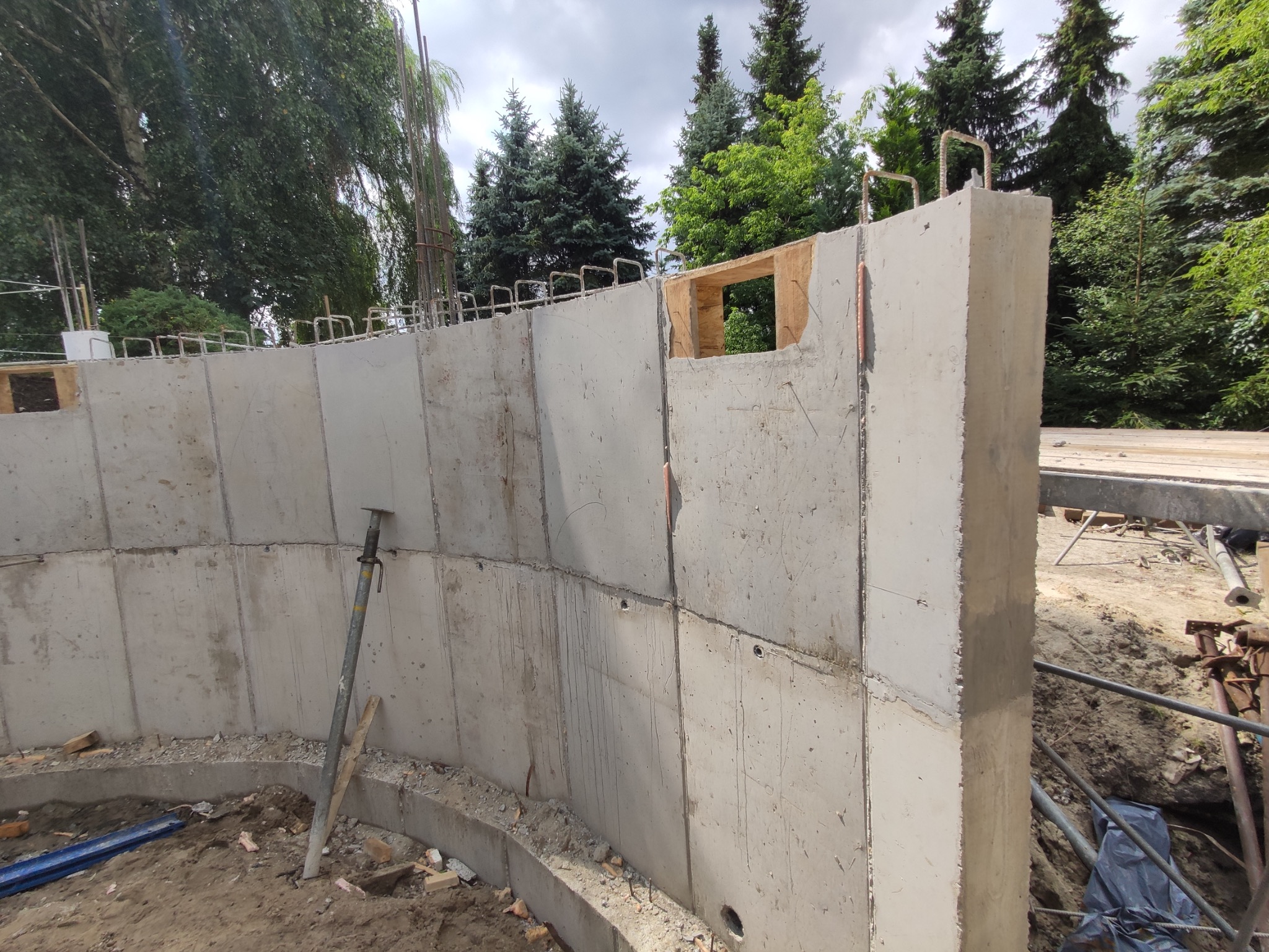 Betonowa ściana fundamentowa w trakcie budowy, widoczne zbrojenie, otwór okienny z drewnianym szalunkiem, podpora stalowa, ziemia i częściowo zarys zieleni w tle.