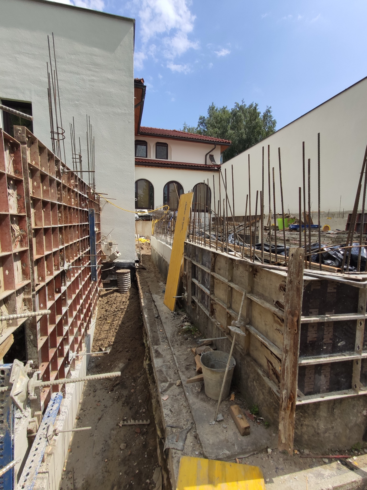 Szczegółowe ujęcie szalunków fundamentowych z wystającymi prętami zbrojeniowymi, widoczne elementy konstrukcyjne i przygotowanie do zalewania betonem, w tle budynek w stanie surowym.