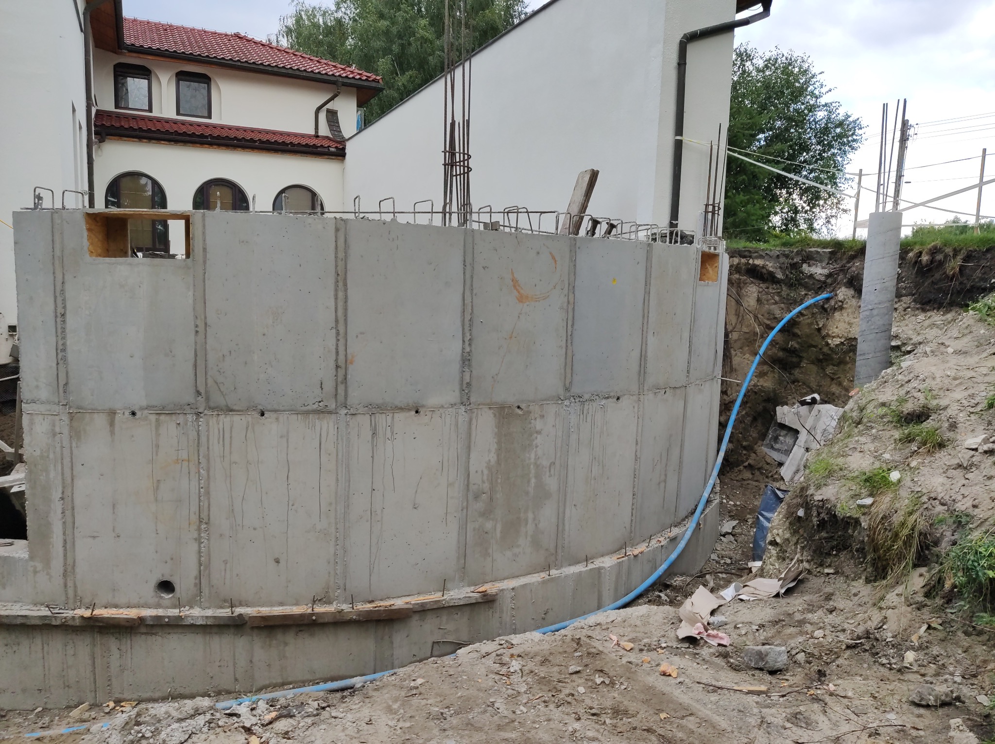 Betonowy fundament budynku w trakcie budowy, widoczne zbrojenia i otwory technologiczne, niebieski przewód w wykopie, w tle biały budynek z czerwonym dachem.