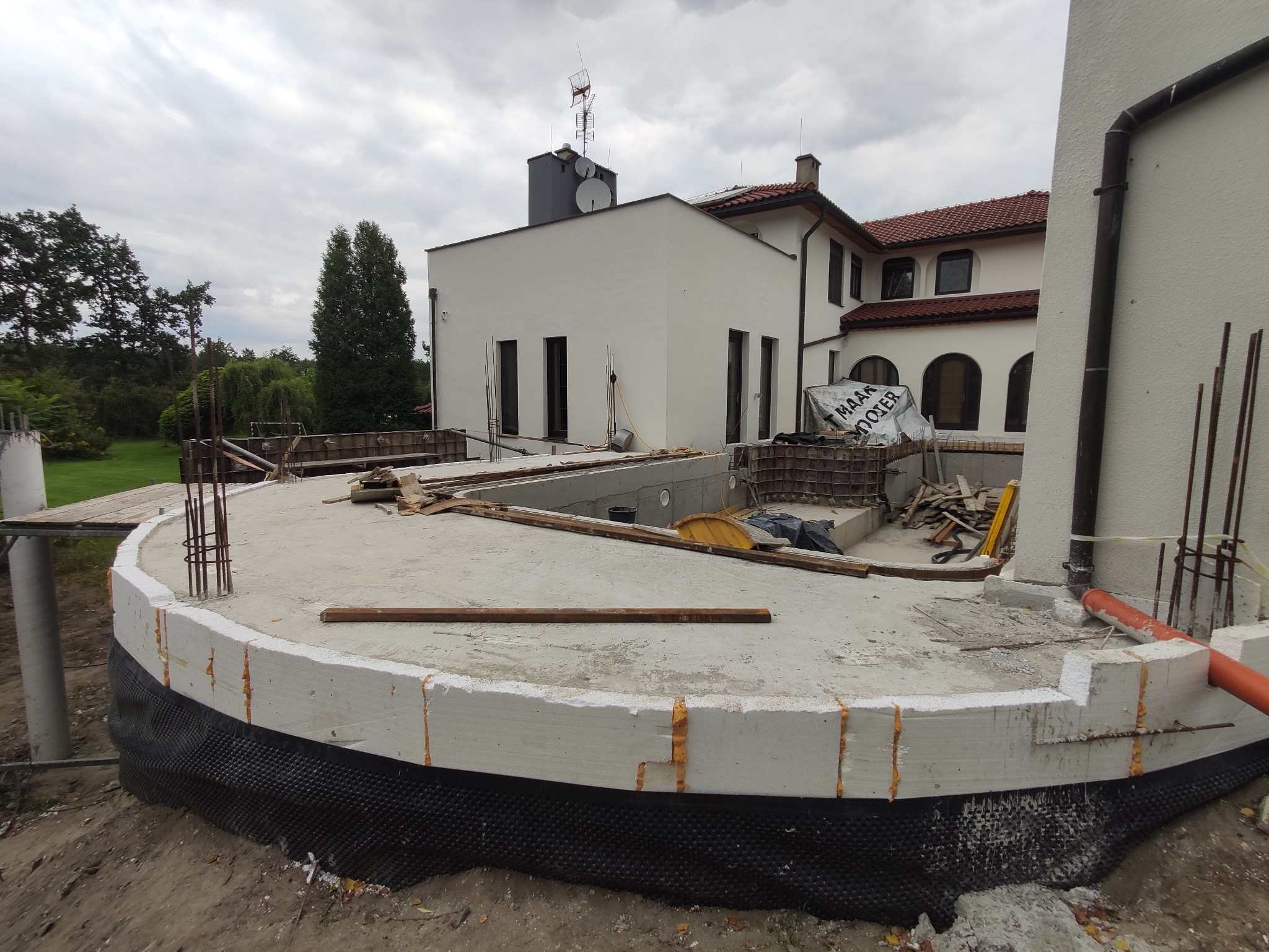 Budowa okrągłego fundamentu domu z widoczną izolacją przeciwwilgociową i wystającymi prętami zbrojeniowymi, na tle częściowo wykończonego budynku w stylu dworkowym.