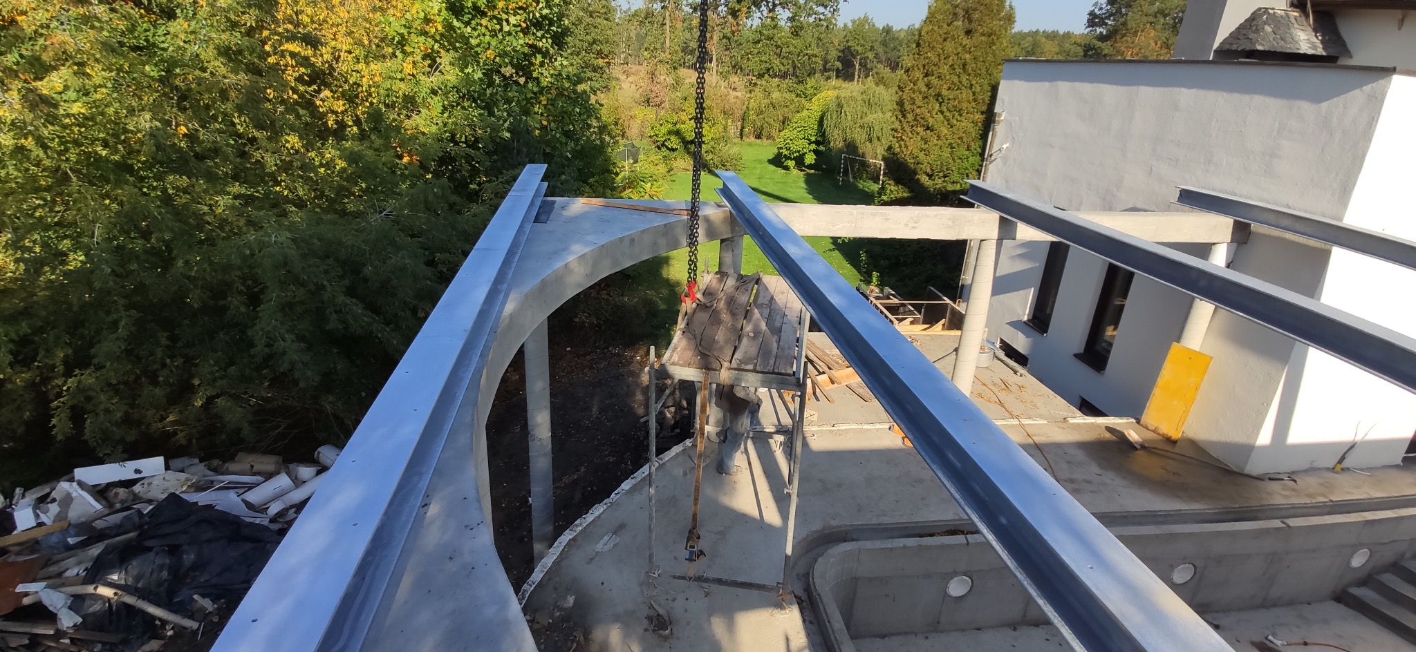 Budowa domu w stanie surowym otwartym, widoczne betonowe elementy konstrukcyjne i stalowe belki, rusztowanie robocze oraz otoczenie z zielenią.