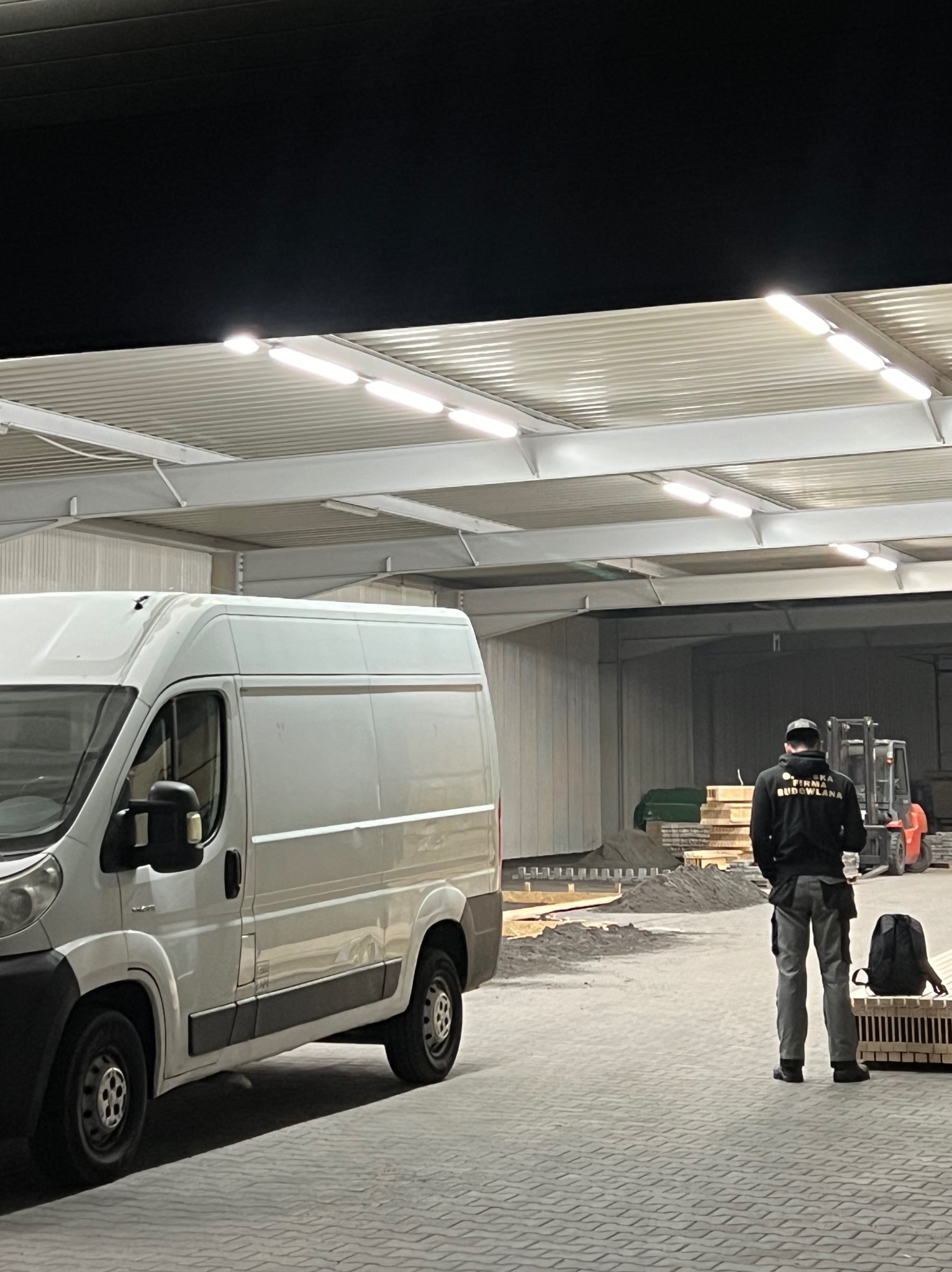 Wnętrze hali z zaparkowanym białym busem, stertami materiałów budowlanych, wózkiem widłowym w tle oraz pracownikiem budowlanym w bluzie z napisem 'Firma Budowlana'.