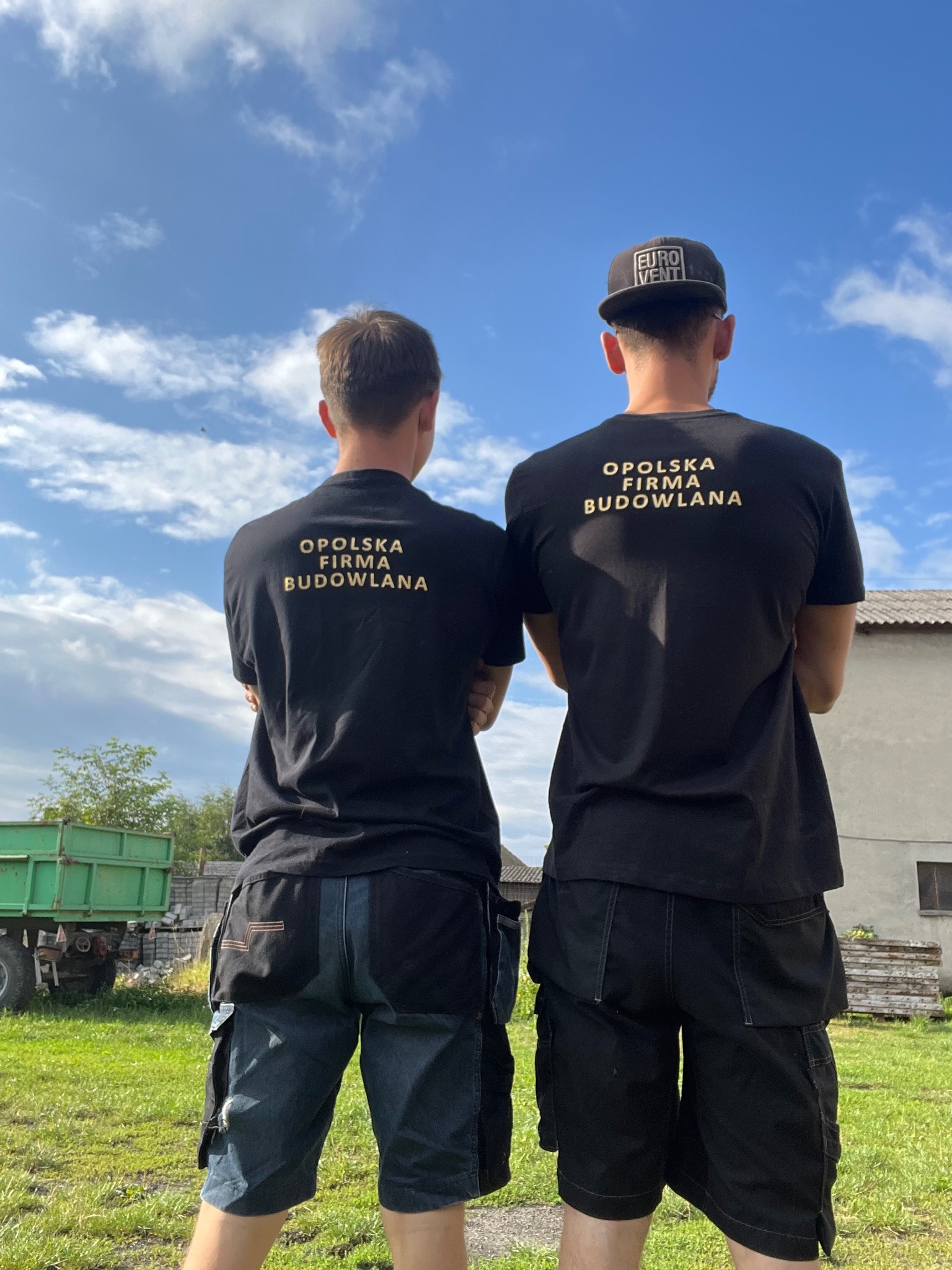 Dwóch mężczyzn w czarnych koszulkach z napisem 'Opolska Firma Budowlana' stoi tyłem, jeden w czapce z daszkiem, drugi w spodniach roboczych z narzędziami, na tle zielonego trawnika, przyczepy...