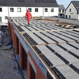 Oleksandr Chema - Budowa domu: strop Teriva zbrojony stalą, robotnik w czerwonym kombinezonie na tle białych domów z panelami słonecznymi. Widok z góry na plac budowy.