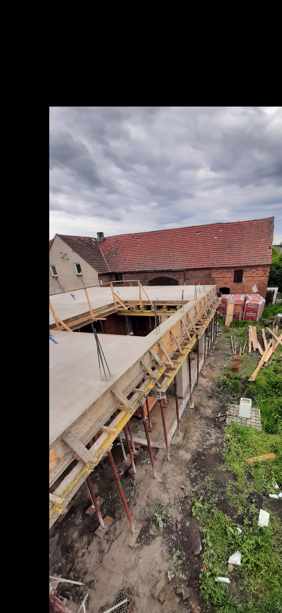 Wylany strop budynku w trakcie budowy, widoczne szalunki, podpory i zbrojenie. W tle budynek z czerwoną dachówką i zachmurzone niebo.