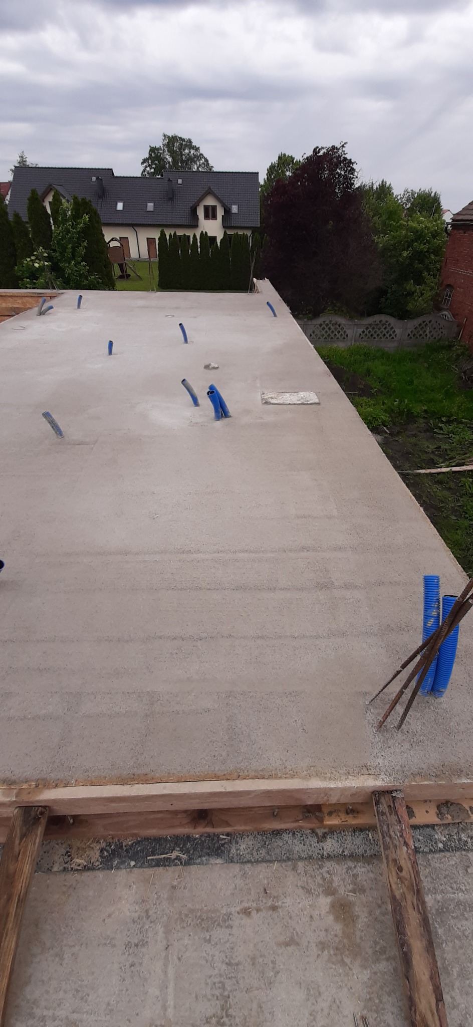 Płyta fundamentowa z wystającymi rurami instalacyjnymi na tle zabudowy jednorodzinnej i pochmurnego nieba. Widok z góry na gotową konstrukcję.