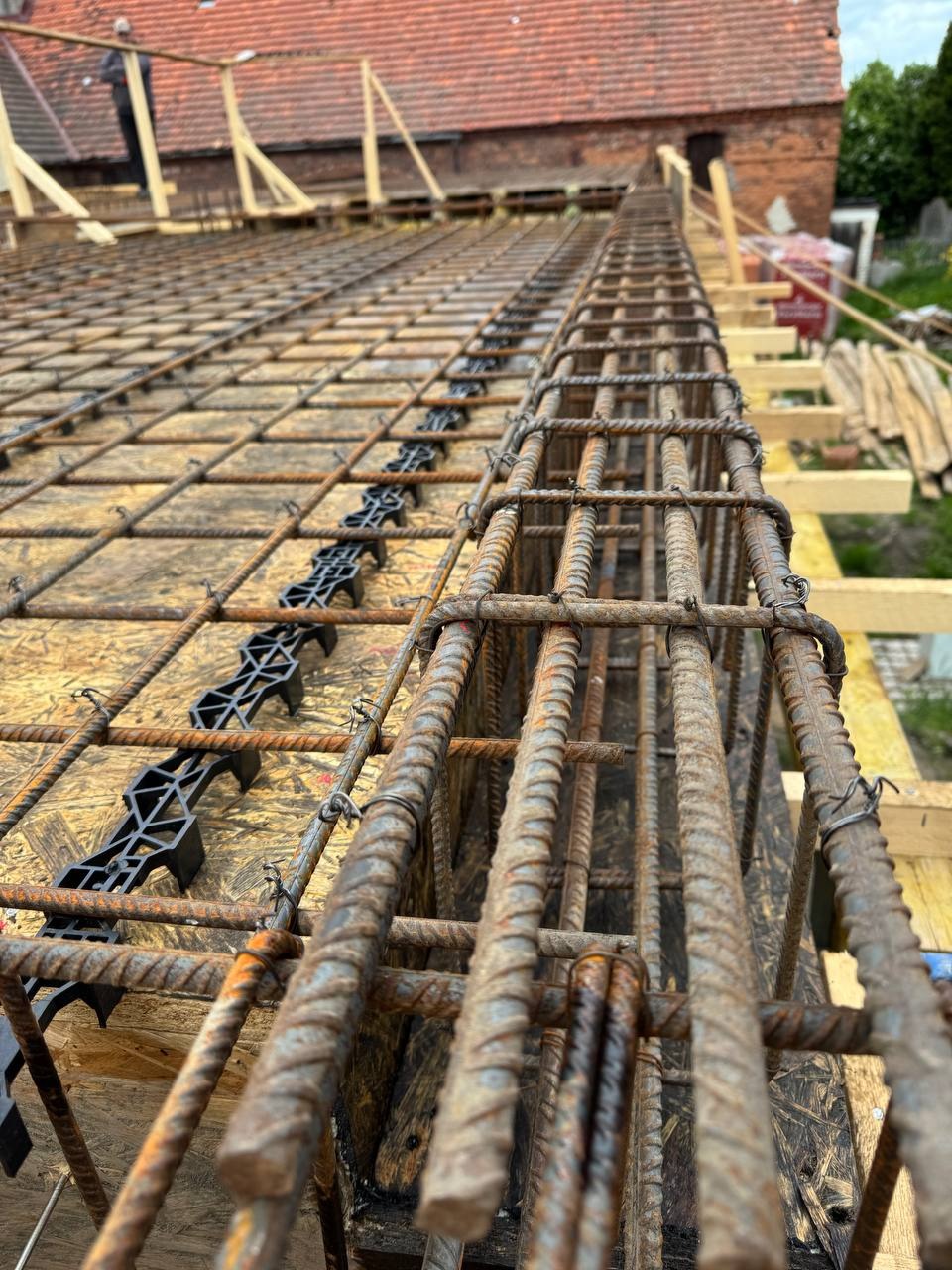 Zbrojenie stropu z widocznymi dystansami z tworzywa, przygotowane do zalania betonem. Widok z dołu na konstrukcję szkieletową i fragment dachu budynku w tle.